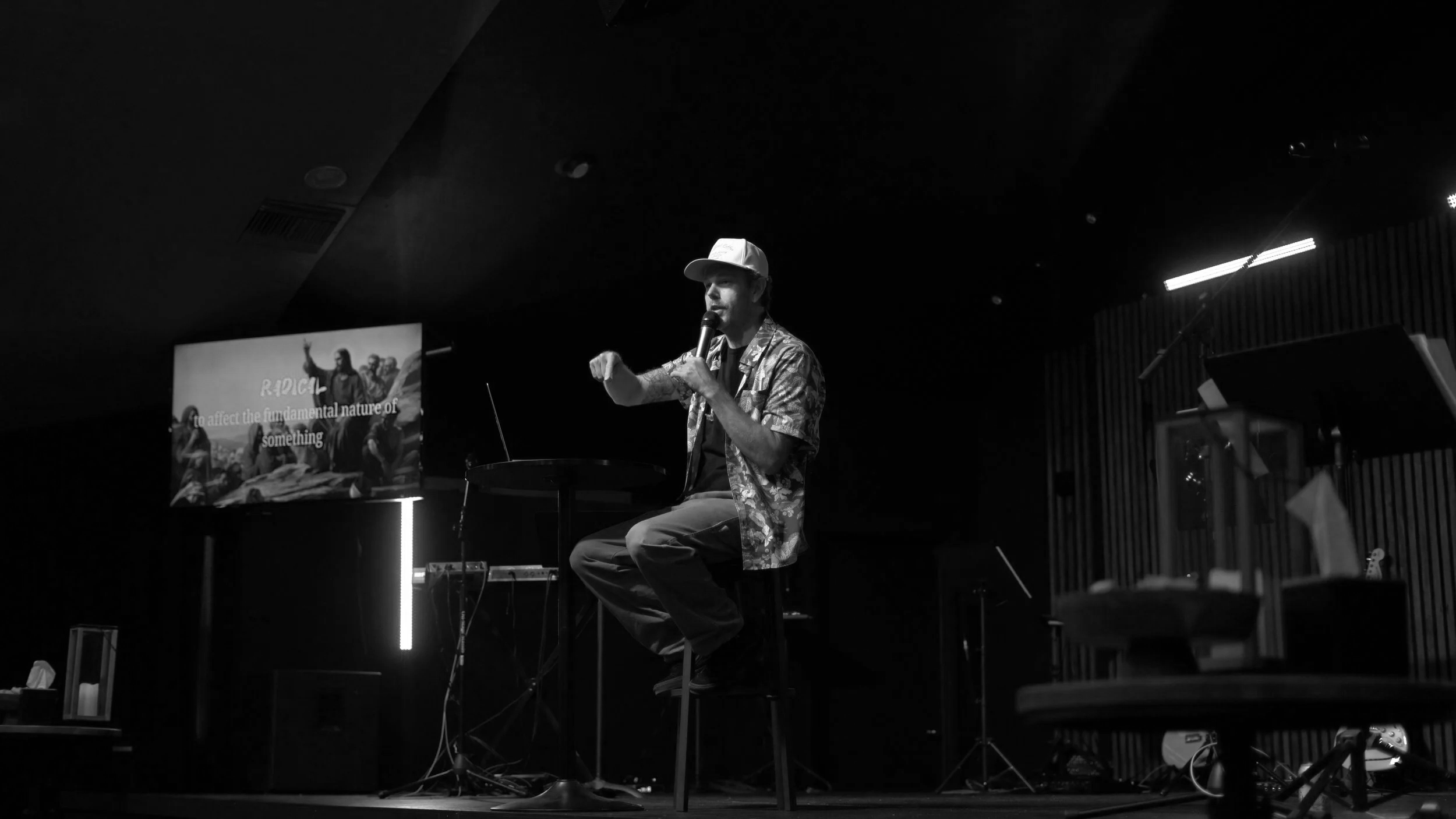 A man wearing a baseball cap and patterned shirt is sitting on a stool on stage, speaking into a microphone during a presentation or performance. There is a large screen behind him displaying a slide with text and images.