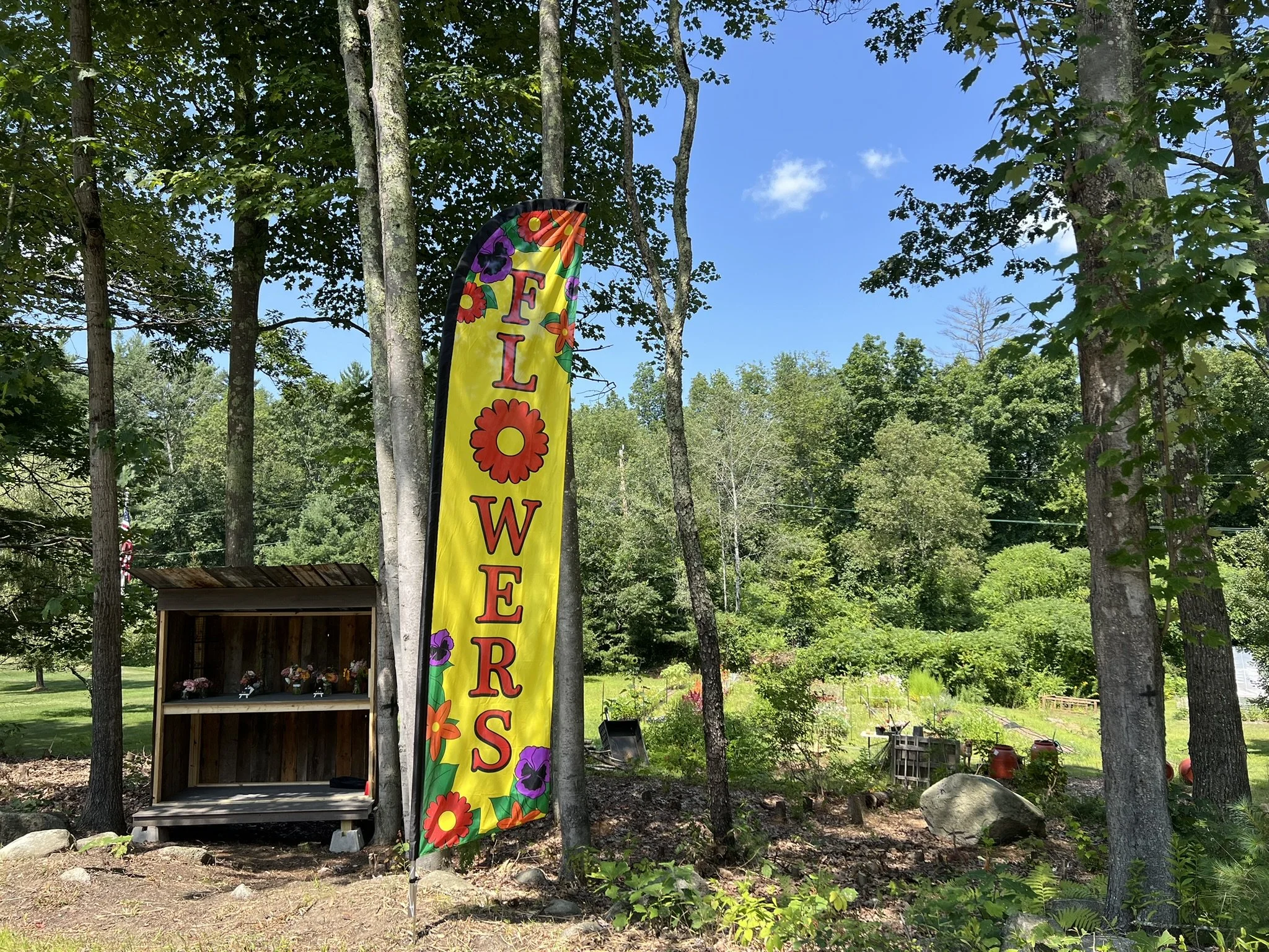 A colorful yellow vertical banner with flowers and the words "FLOWERS" in pink letters, set outdoors among trees, with a small wooden stand and garden supplies below.