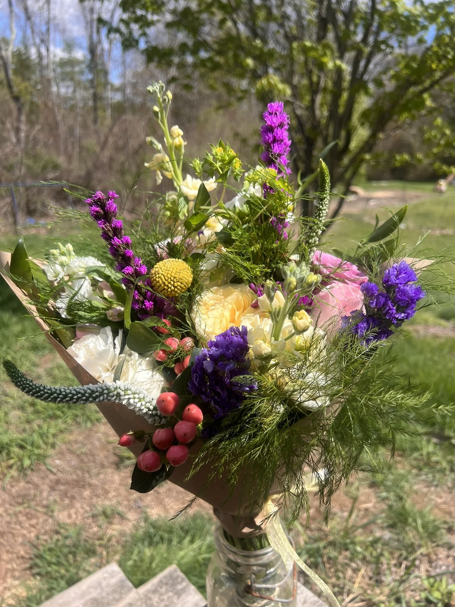 A bouquet of colorful mixed flowers held in a glass jar outdoors with trees and grass in the background.