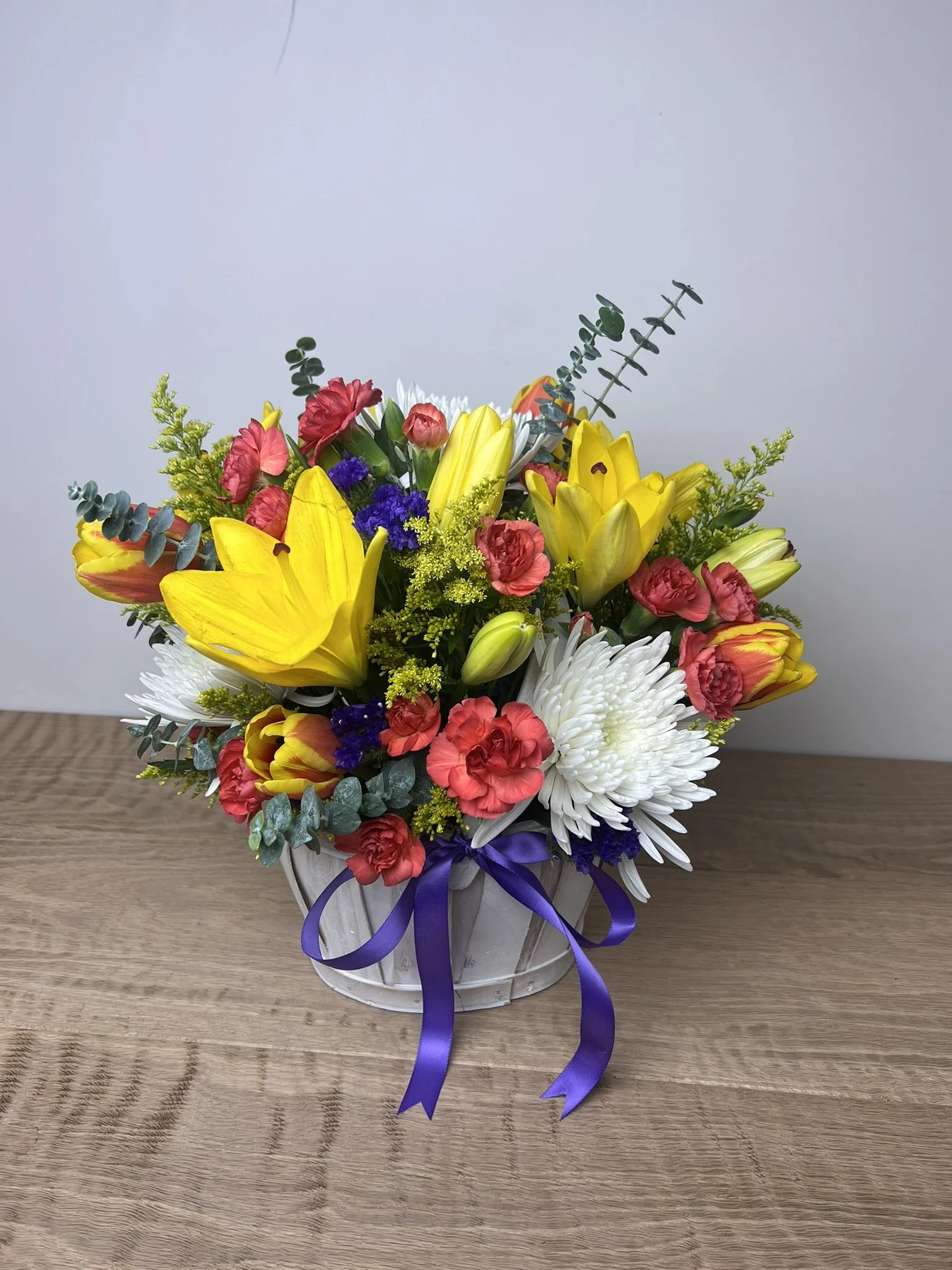A colorful bouquet of flowers in a white basket with a purple ribbon, placed on a wooden surface, against a plain light-colored wall.