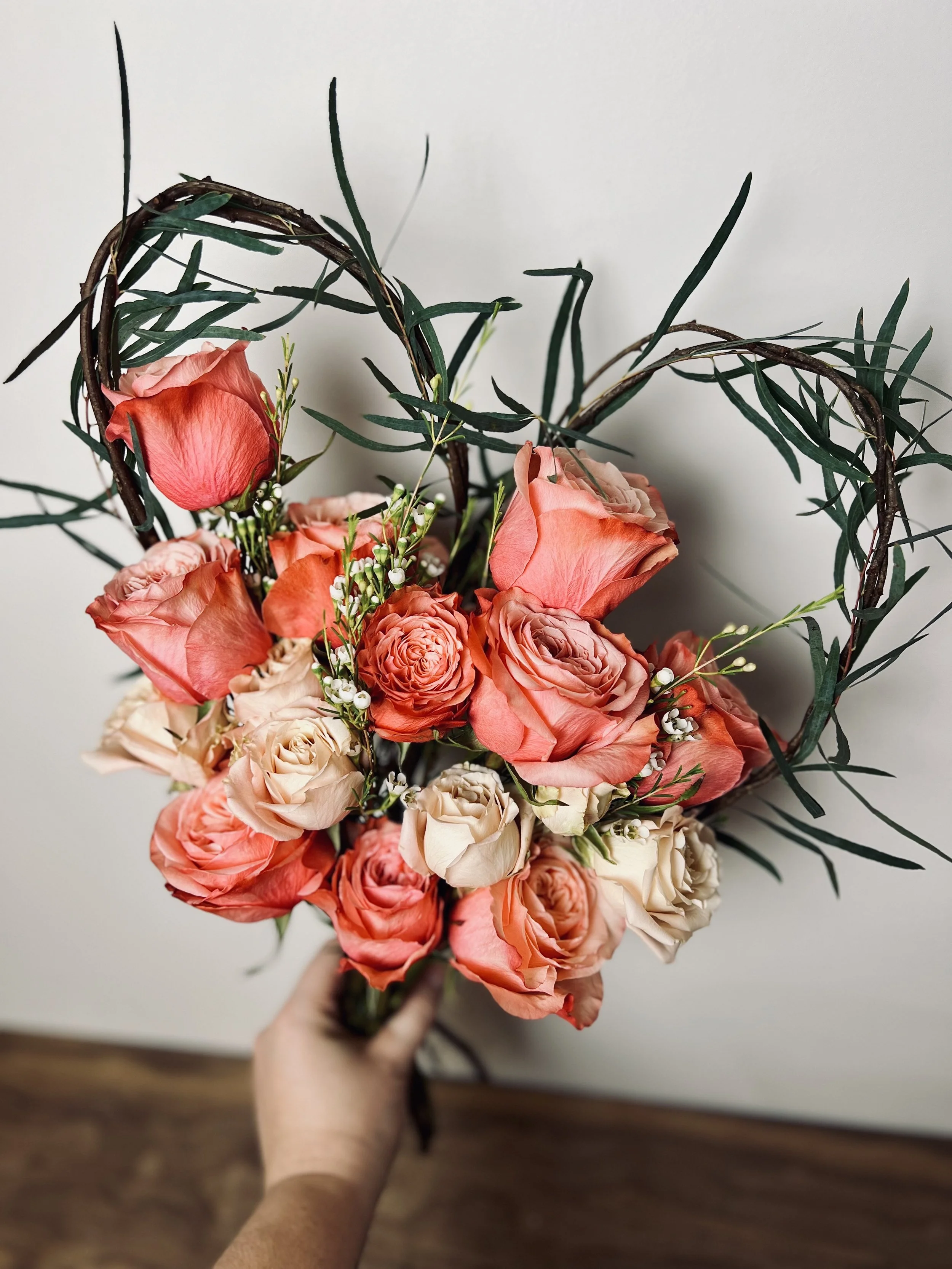 Person holding a bouquet of mixed pink and peach-colored flowers with green leaves and stems, against a plain white wall background.