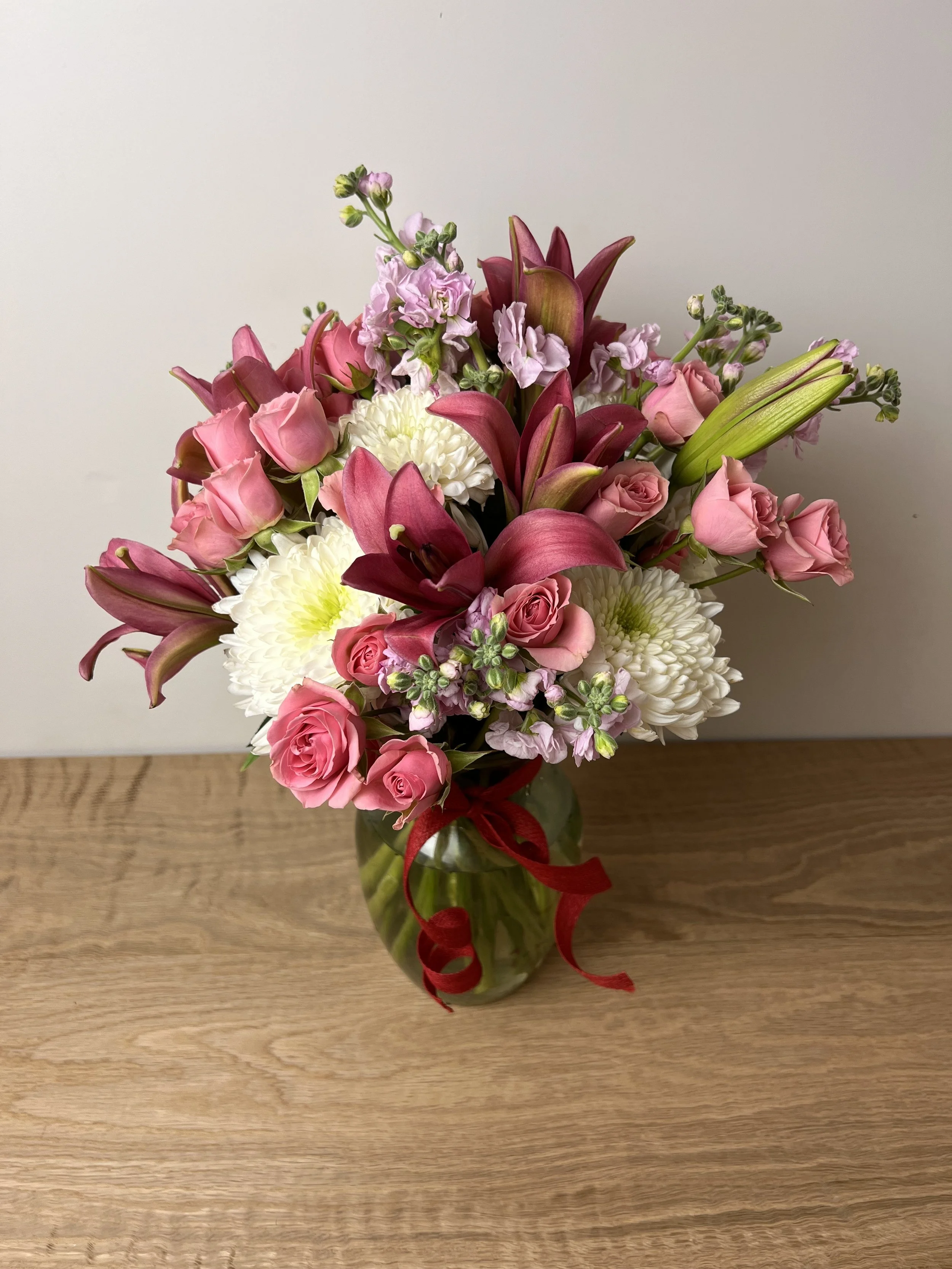 A bouquet of pink roses, white chrysanthemums, pink stock flowers, and deep red lilies in a glass vase tied with a red ribbon, placed on a wooden surface against a plain background.