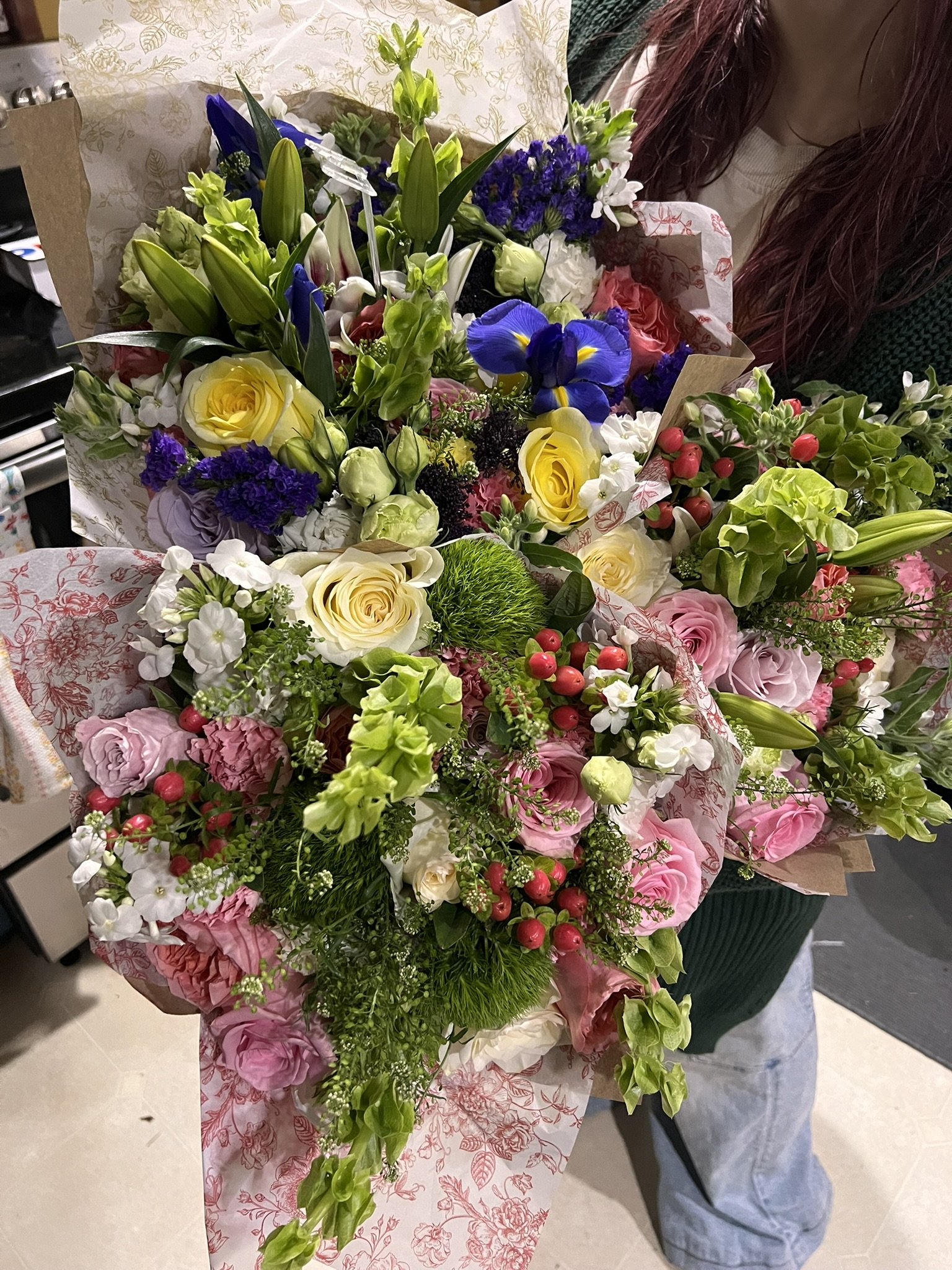 A large colorful bouquet of flowers including yellow roses, white flowers, purple irises, pink roses, red berries, and greenery held by a person wearing a green sweater and gray apron.