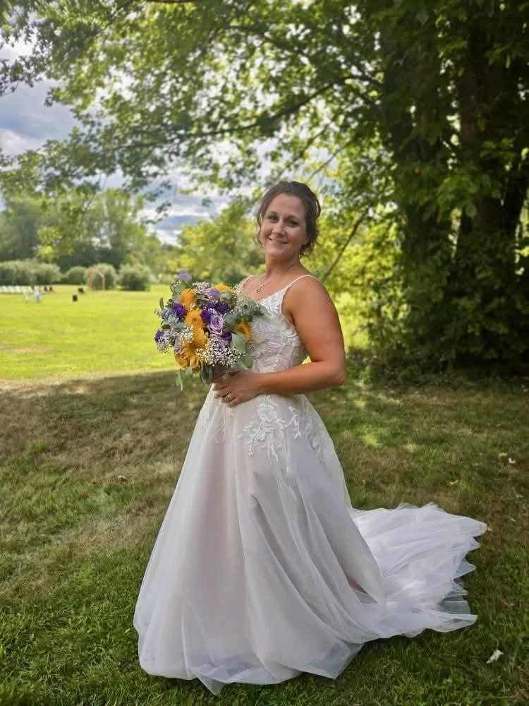 A woman in a wedding dress holding a bouquet of colorful flowers, standing outdoors on grass with trees and a cloudy sky in the background.
