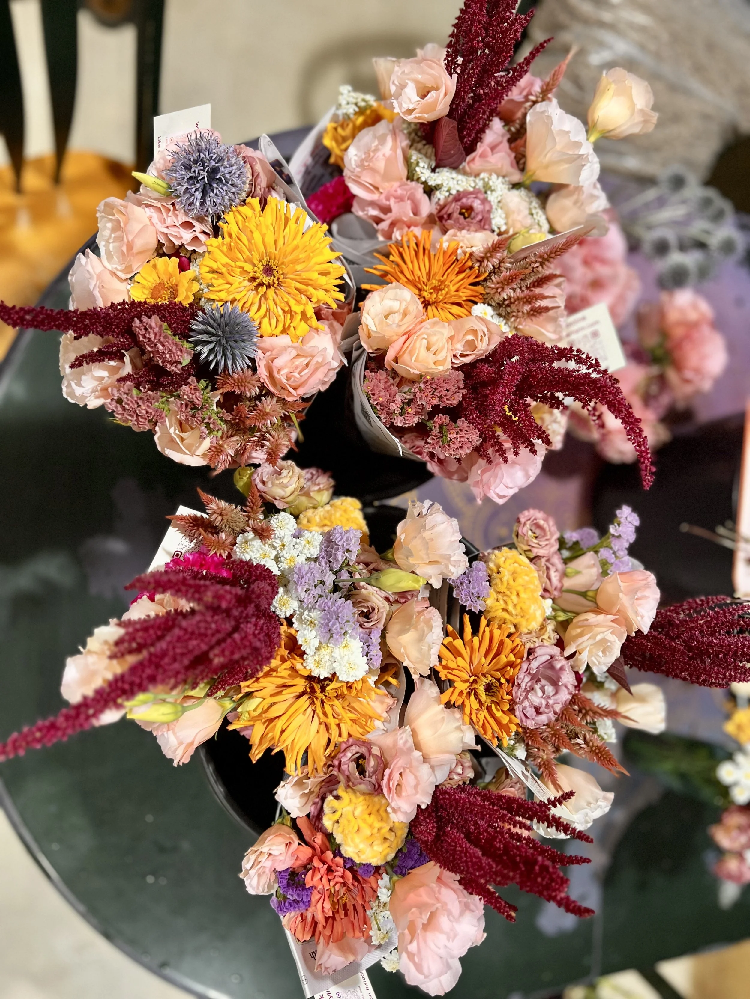 A colorful bouquet of mixed flowers including pink roses, orange marigolds, purple globe amaranth, red amaranth, and white baby's breath, arranged on a black circular surface.