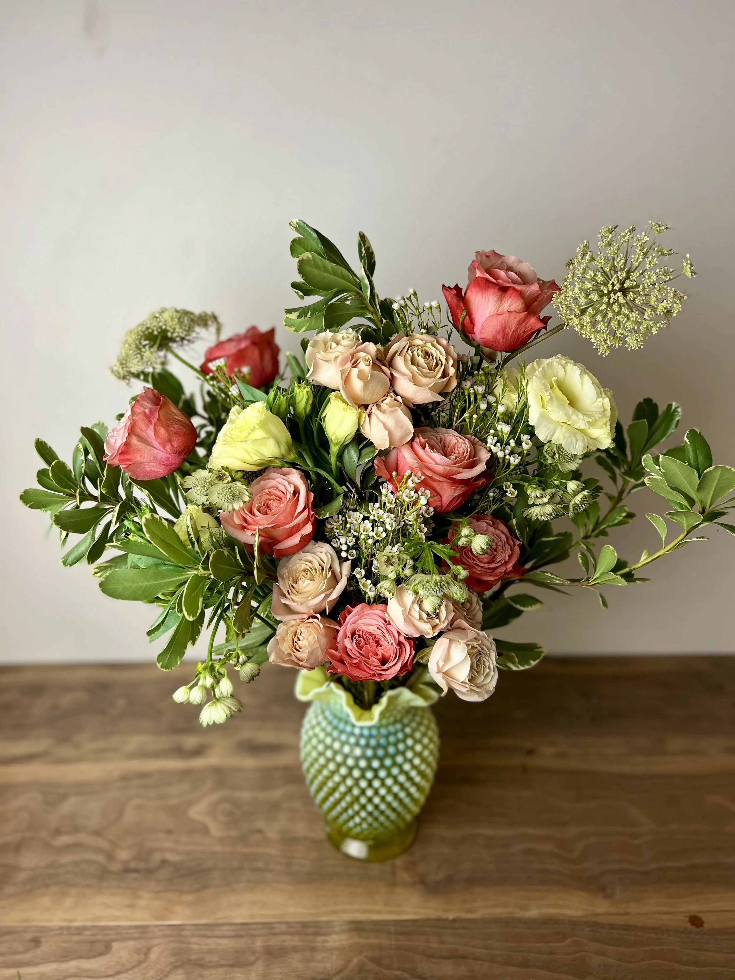 A bouquet of pink, red, and cream roses with greenery and white filler flowers in a green textured vase on a wooden table.
