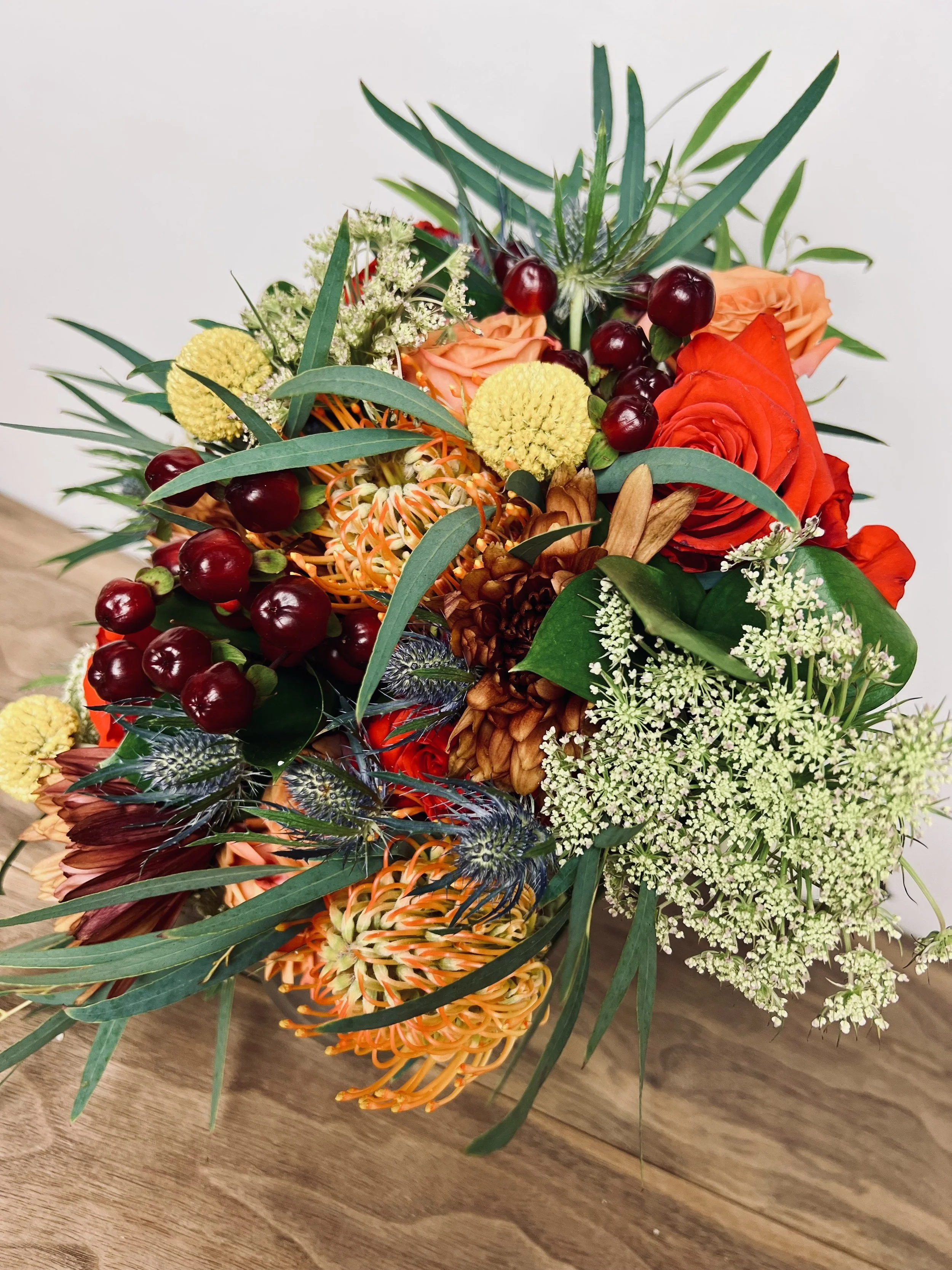 A colorful bouquet of flowers including roses, berries, and various greenery, arranged on a wooden surface.