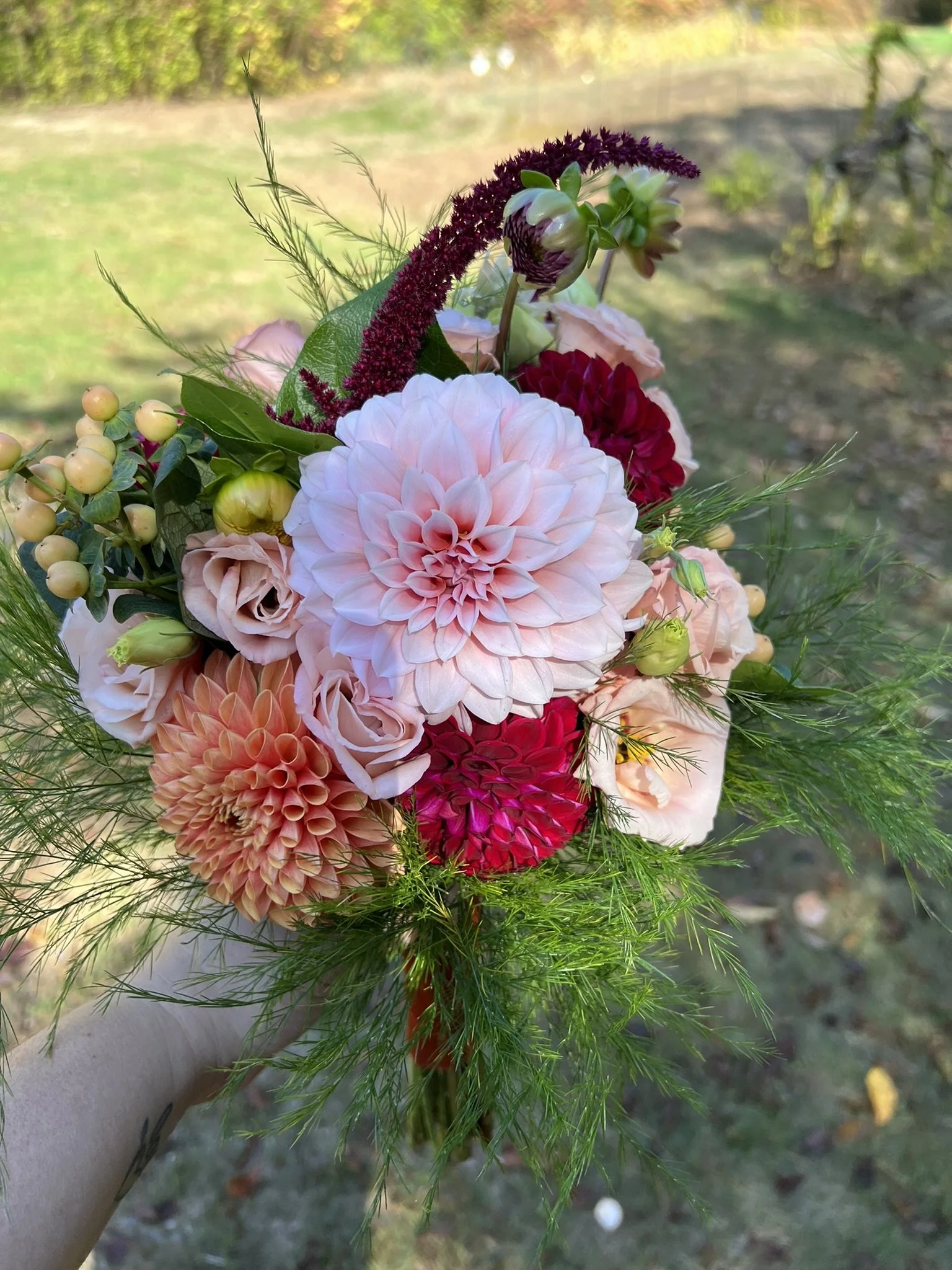 A colorful bouquet of flowers with blush pink, red, and burgundy flowers, accented with green leaves and fern-like greenery, held outdoors with a blurred natural background.