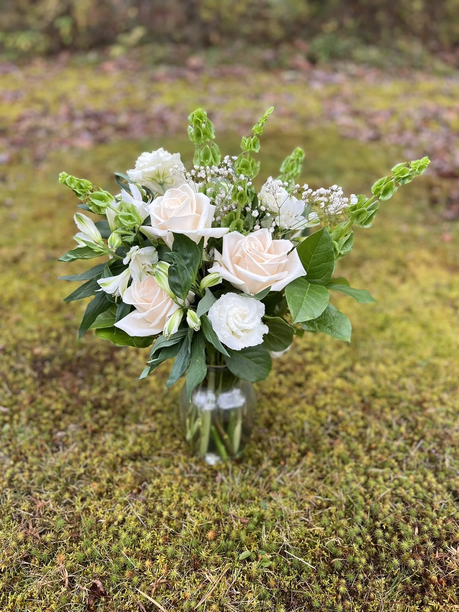 A bouquet of white and cream roses, white lisianthus, and small white baby's breath flowers with lush green leaves, arranged in a clear glass vase on a moss-covered ground outdoors.