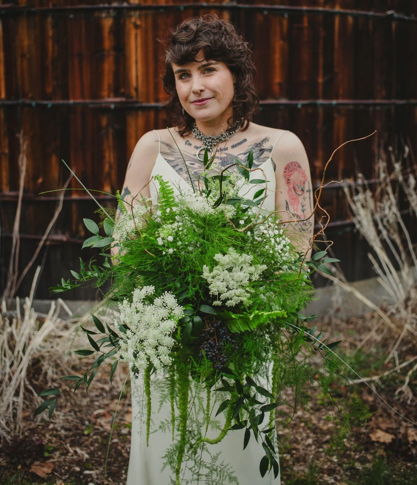 Woman with short curly hair holding a large bouquet of green and white plants and flowers outdoors in front of a wooden fence.