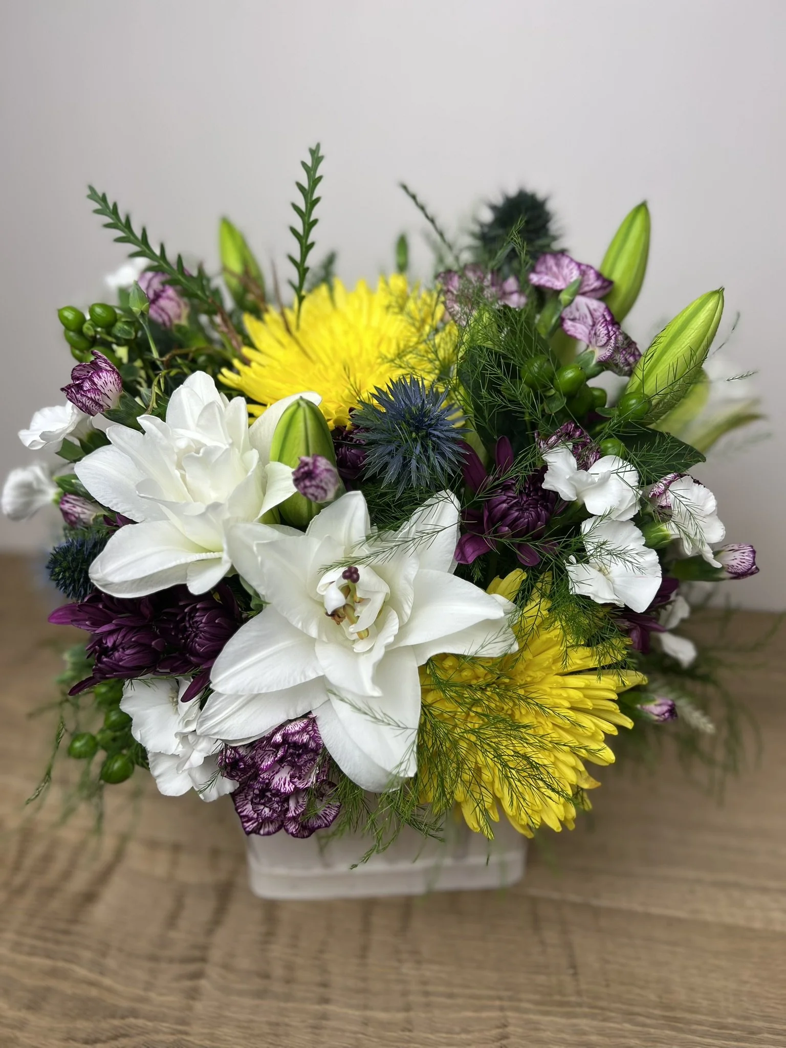 A colorful bouquet of various flowers, including white lilies, purple chrysanthemums, yellow chrysanthemums, and pink striped flowers, arranged with greenery, placed on a wooden surface.