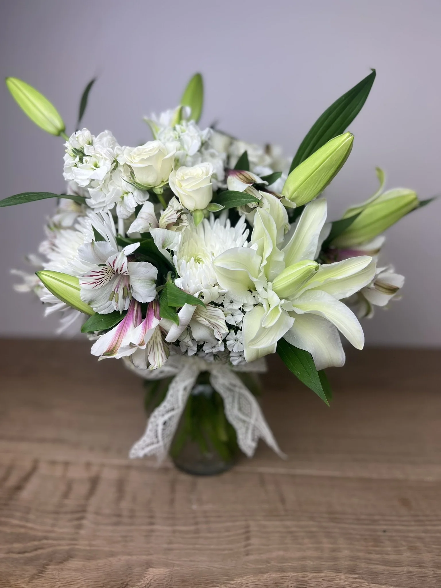 A bouquet of white flowers including lilies, roses, and daisies, tied with a lace ribbon, on a wooden surface.