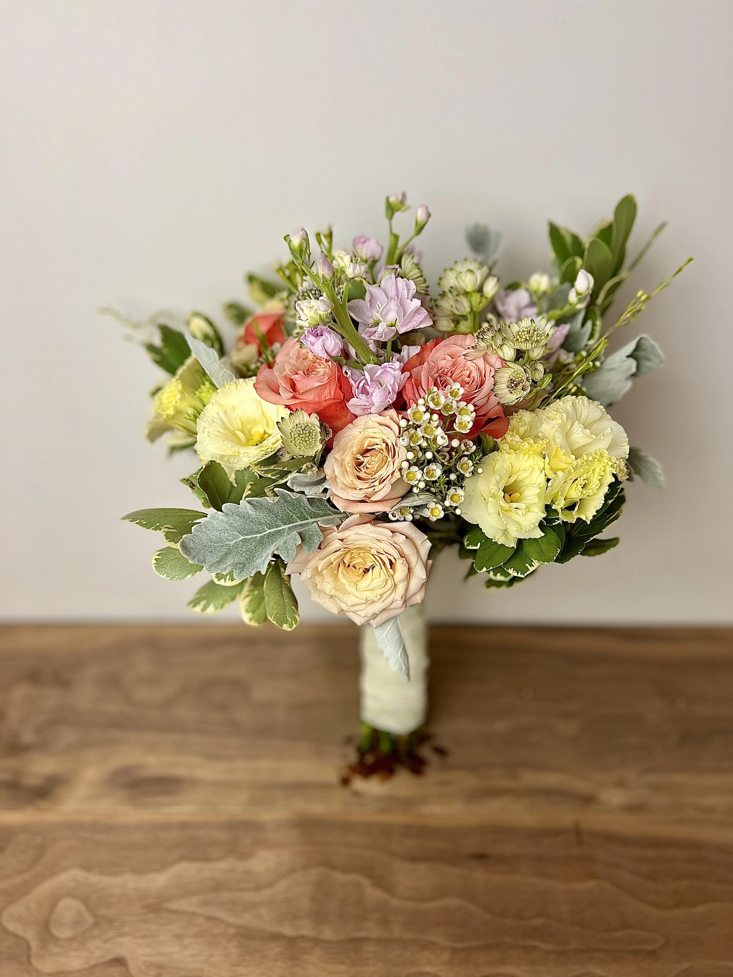 A bouquet of mixed flowers including roses, lisianthus, and various greenery, arranged with a cream-colored ribbon wrapped around the stems, resting on a wooden surface.