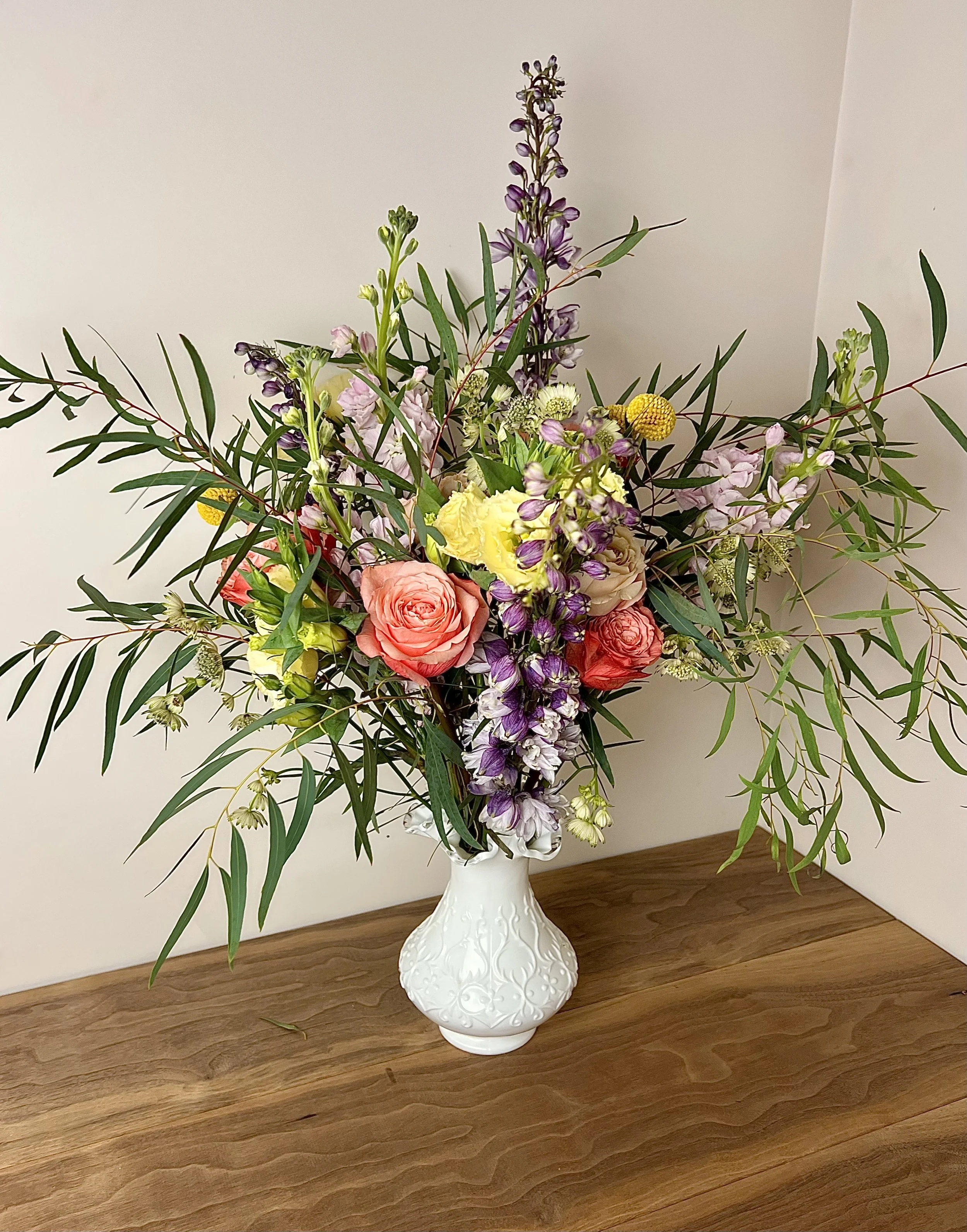 A white ceramic vase with floral design on a wooden table, holding a bouquet of mixed flowers and greenery against a plain white wall.
