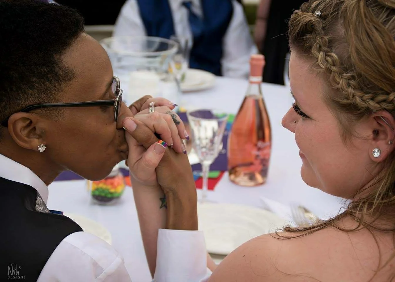 Two women holding hands and touching foreheads during a wedding or celebration, sitting at a decorated table.