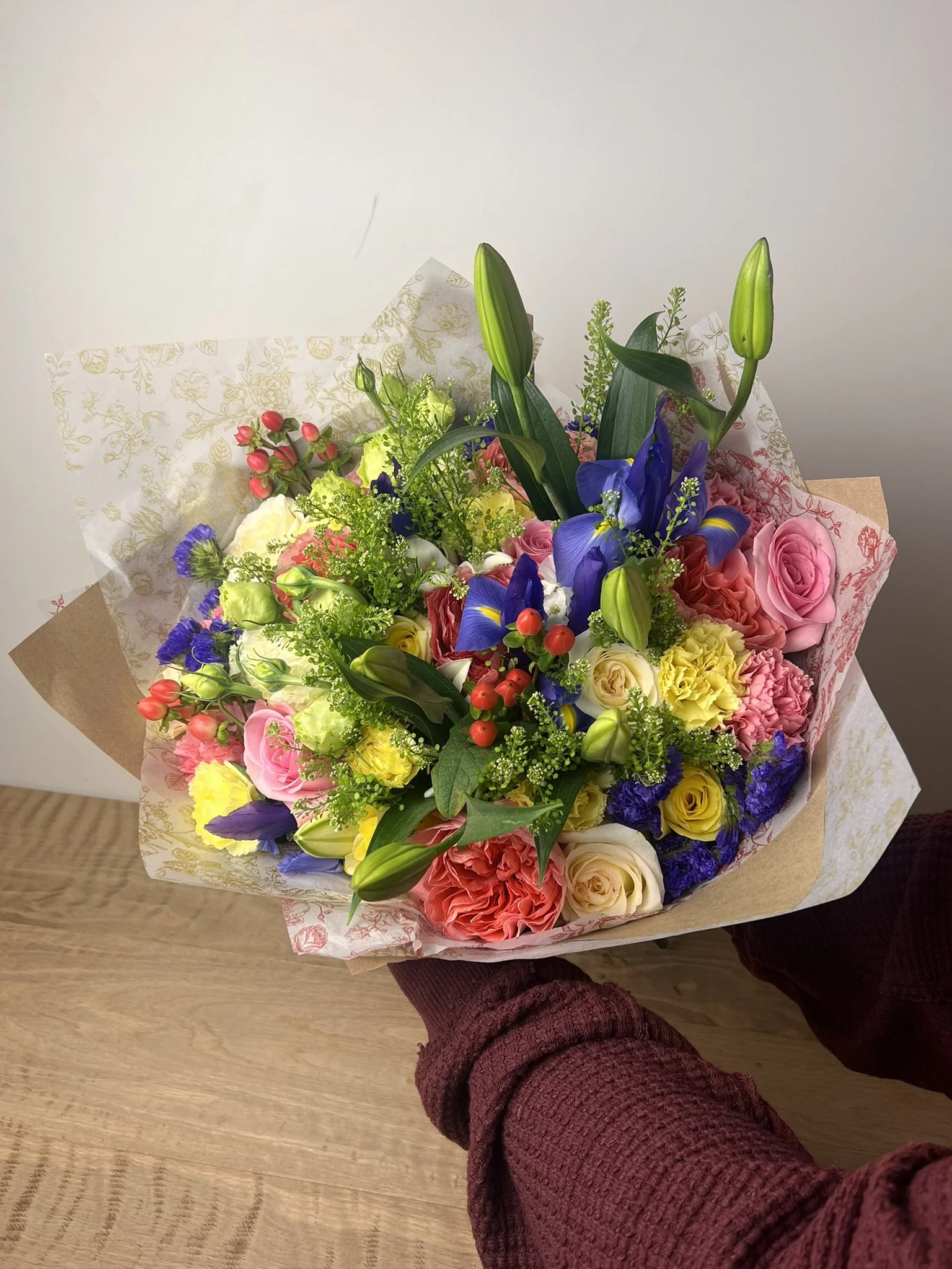Person holding a colorful bouquet of flowers, including pink roses, yellow flowers, purple irises, white roses, and greenery, with a plain white wall in the background.