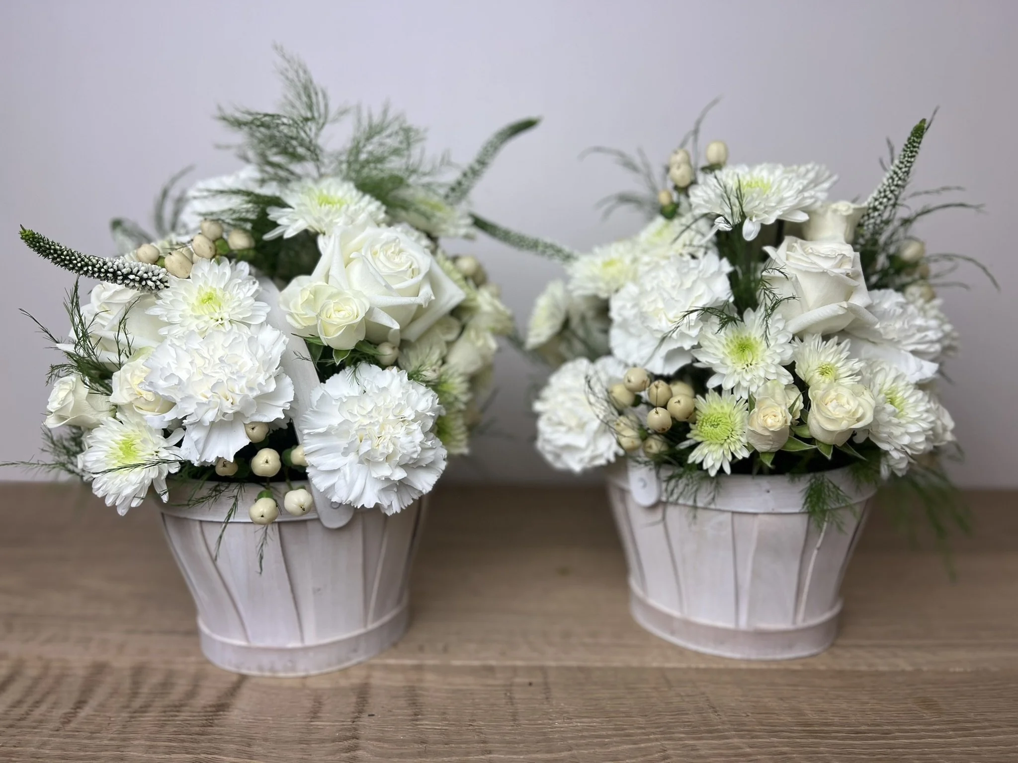 Two white flower arrangements in white vases on a wooden surface.