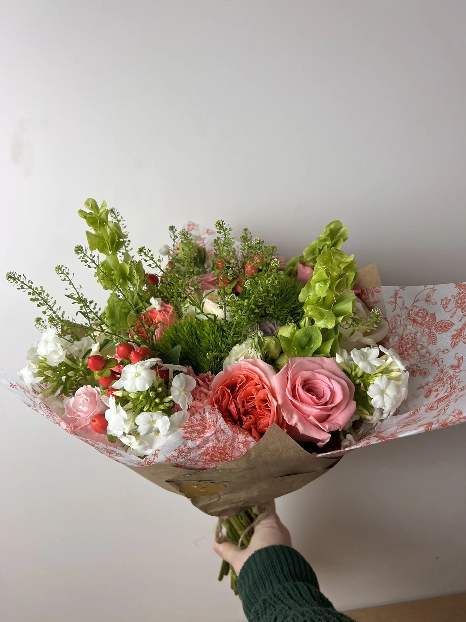 A hand holding a bouquet of mixed flowers including pink roses, white blossoms, green foliage, and red berries, wrapped in decorative paper.