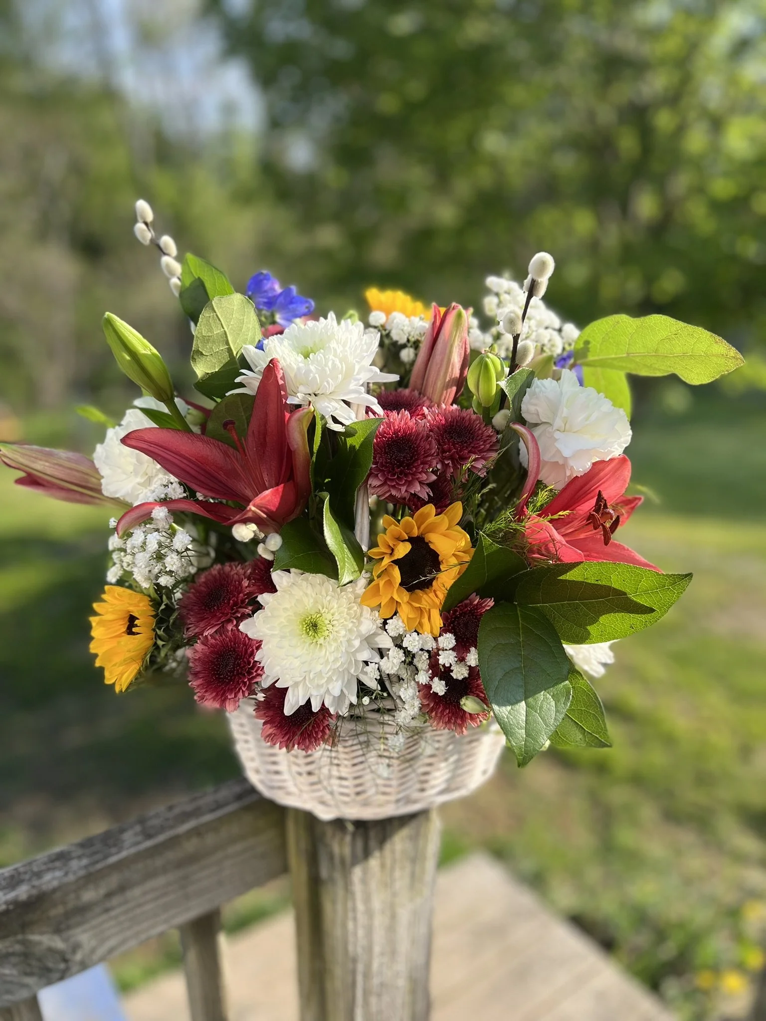 A bouquet of mixed flowers including white daisies, pink lilies, yellow sunflowers, and purple blooms in a white basket on a wooden railing outdoors.