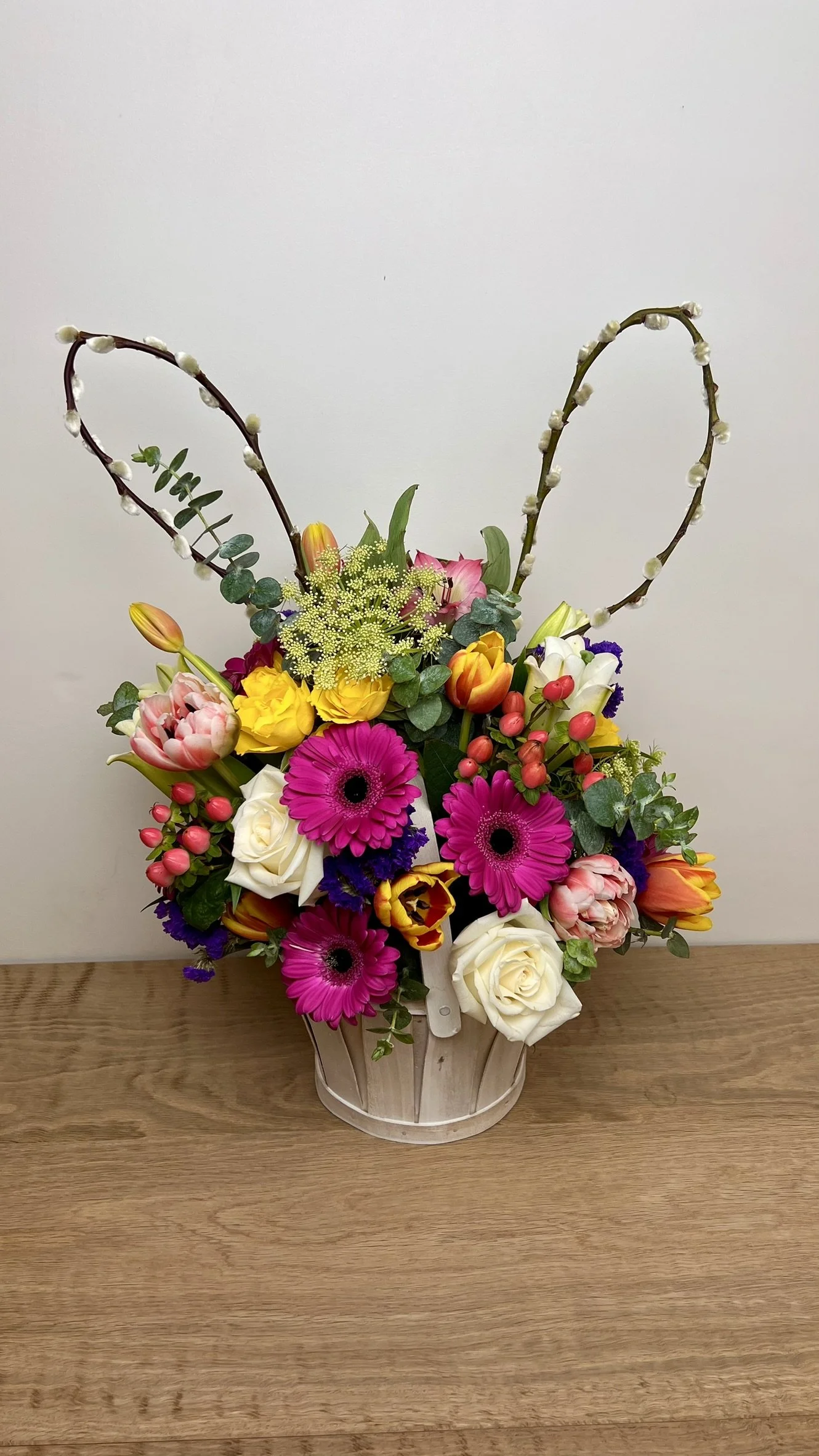 A bouquet of colorful flowers including pink gerbera daisies, white roses, yellow tulips, orange tulips, purple statice, pink hypericum berries, and green foliage in a white wooden basket on a wooden table against a plain white wall.
