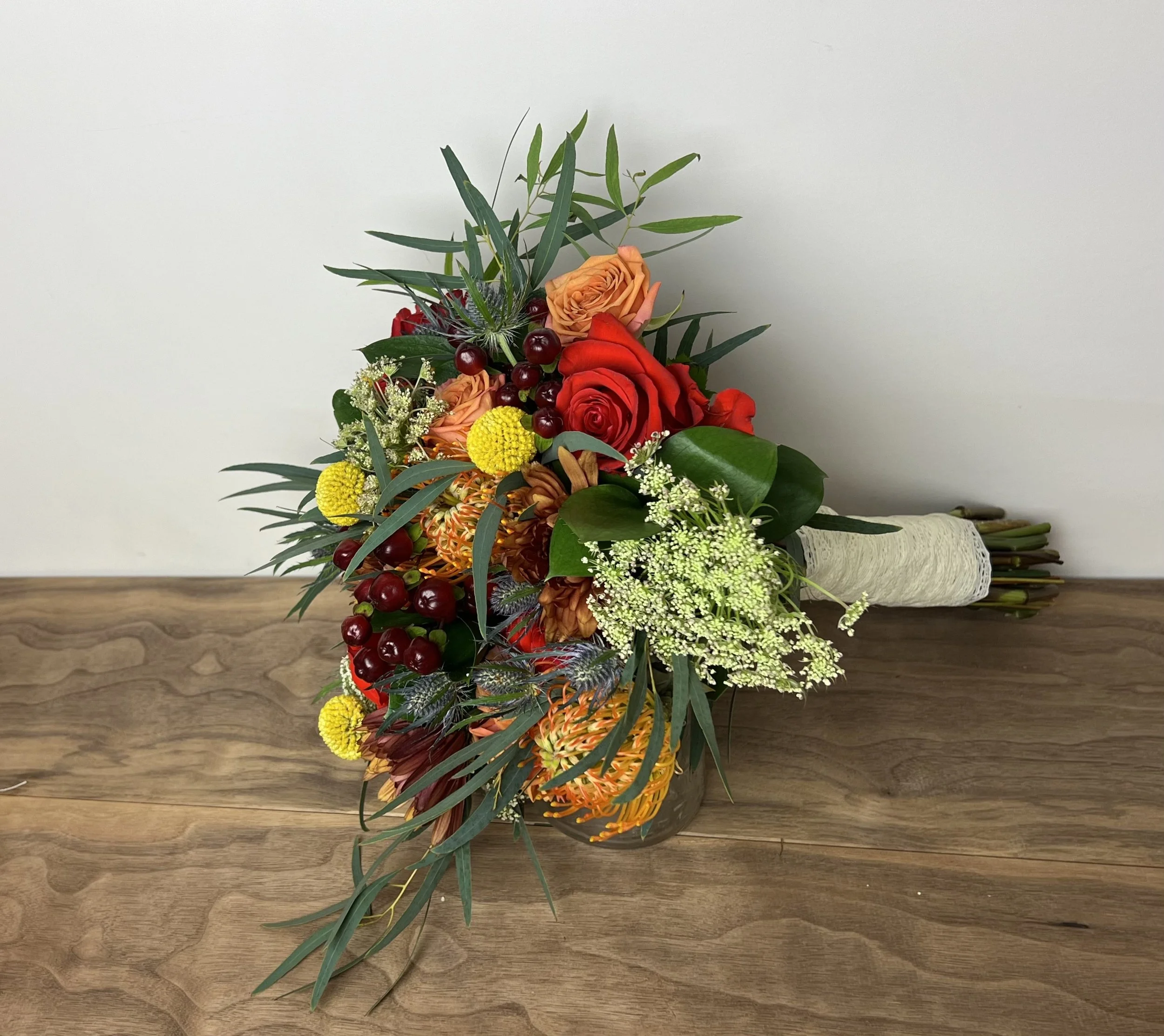 A bouquet of flowers with red, orange, and yellow blooms, green leaves, and small white and red accents, arranged in a clear glass vase on a wooden surface against a plain white background.