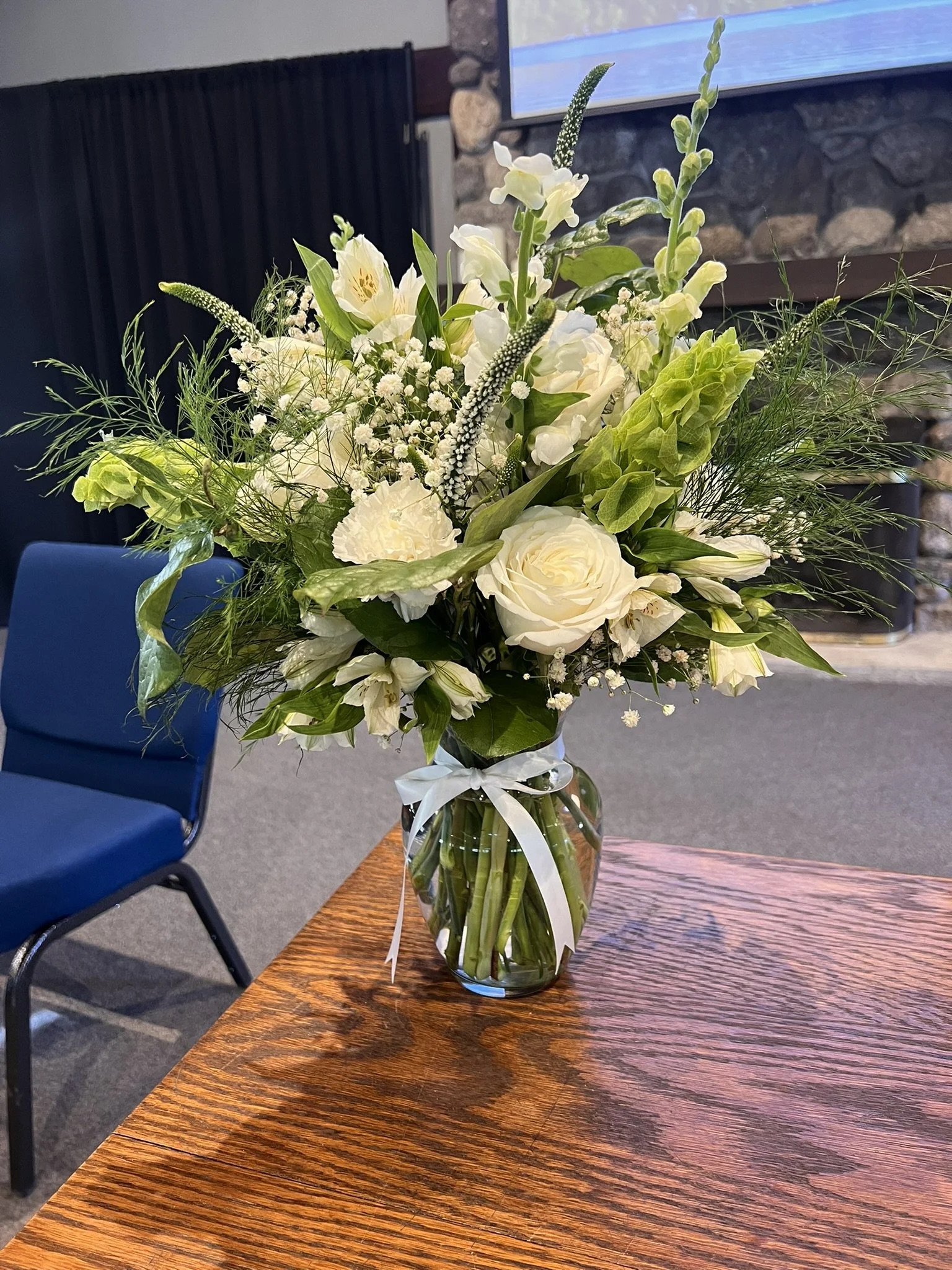 A tall clear glass vase with a white ribbon tied around the neck, holding a bouquet of white and green flowers on a wooden table.