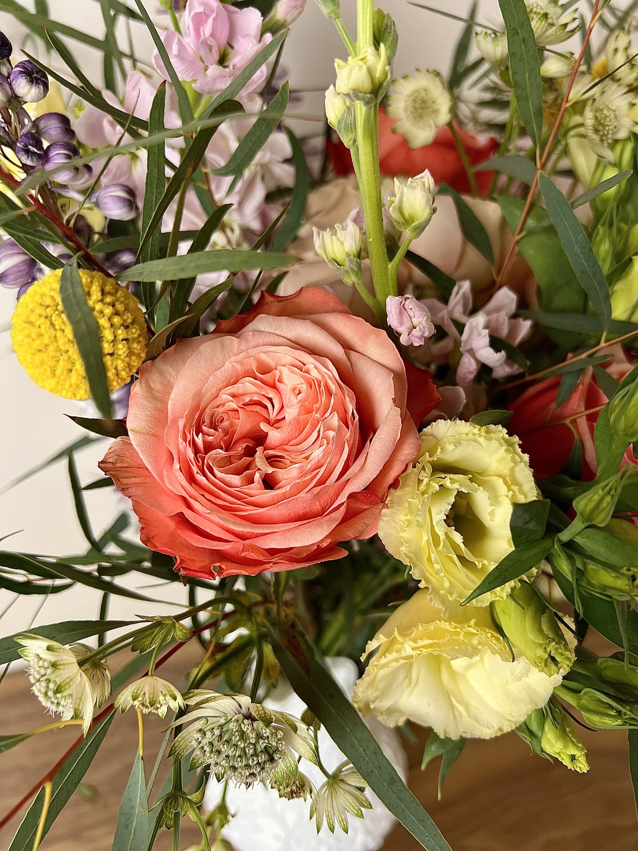 A close-up of a mixed flower bouquet with pink, yellow, peach, green, and purple flowers and green leaves.