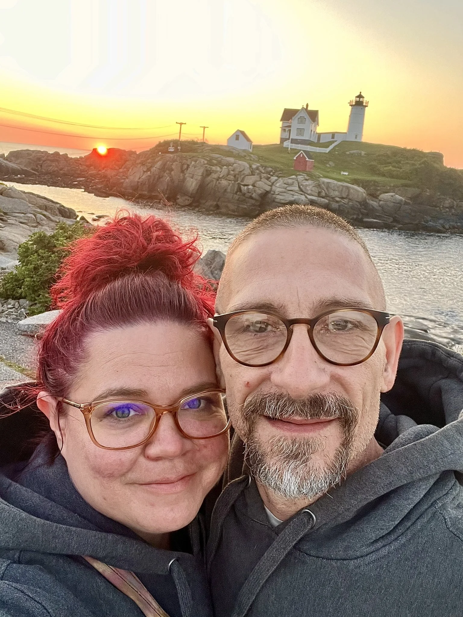 Close-up photo of a couple taking a selfie outdoors during sunset, with a lighthouse and rocky coastline in the background.