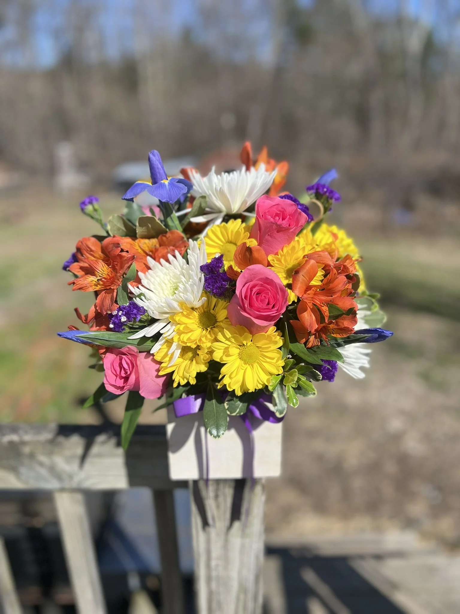 A colorful bouquet of various flowers including pink roses, white daisies, purple irises, and orange lilies, arranged in a narrow white container outdoors.