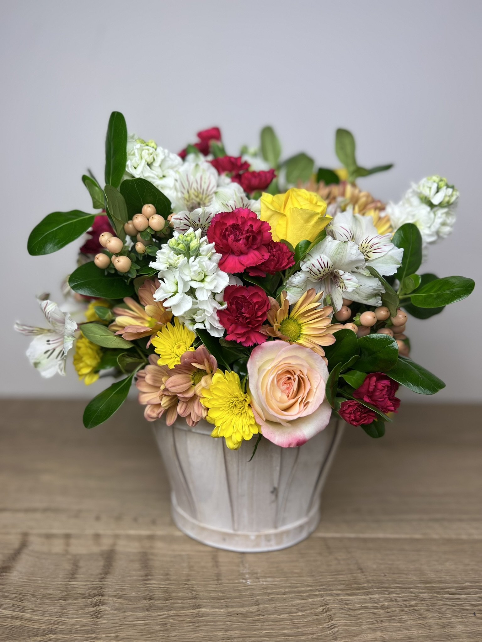 A white ceramic vase filled with colorful flowers including pink carnations, yellow roses, white lilies, yellow daisies, and greenery, placed on a wooden surface against a plain background.
