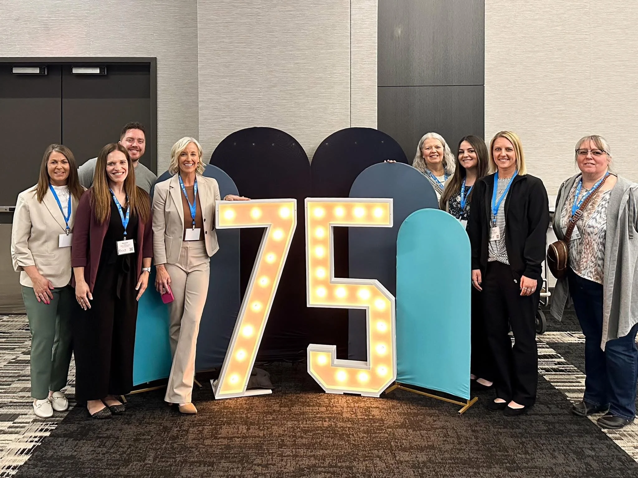 A group of diverse smiling people standing together behind decorative light-up letters and numbers at a conference or event.