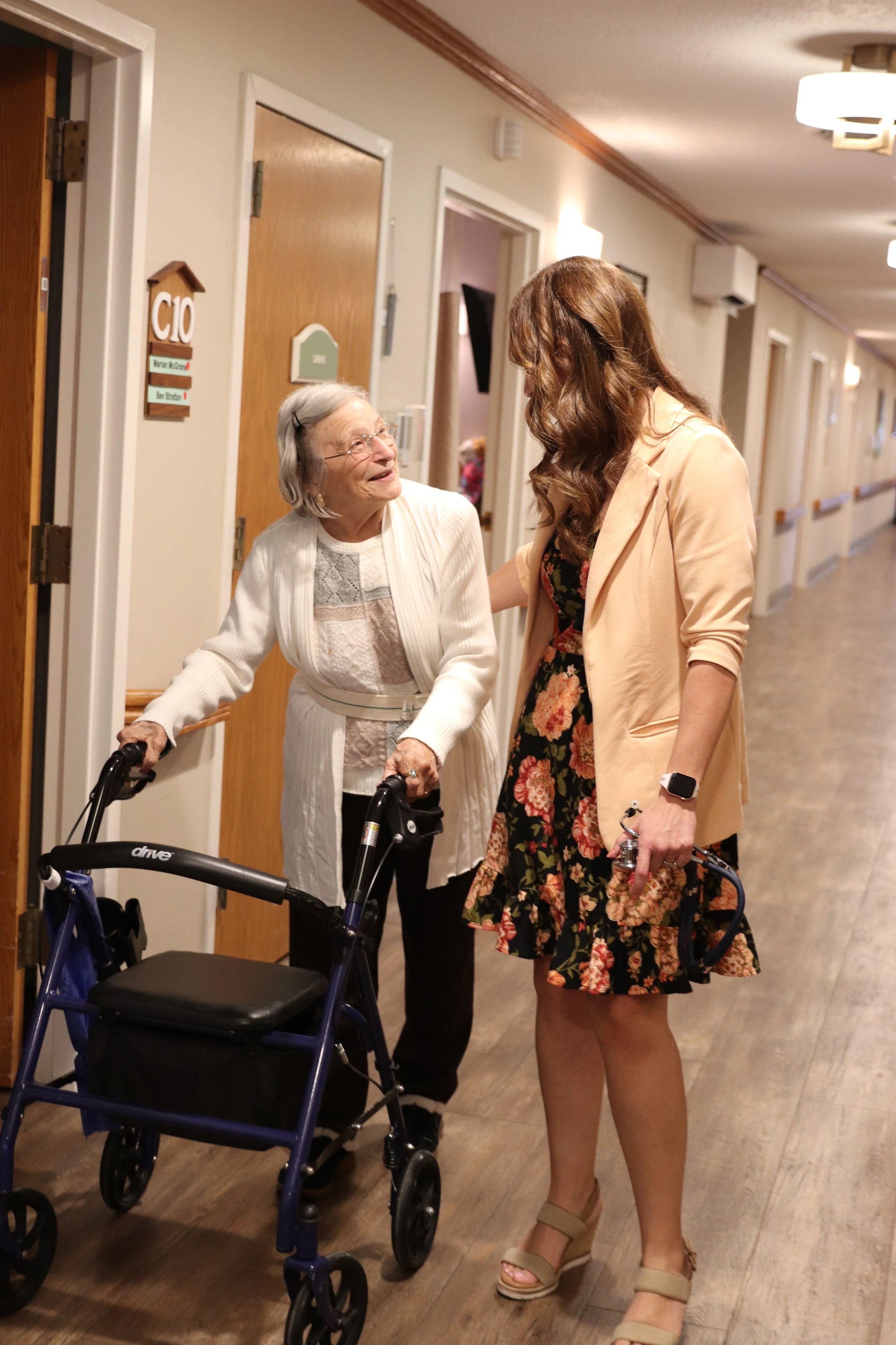 An elderly woman with gray hair using a walker, smiling and talking to a younger woman with long brown hair wearing a floral dress and beige jacket. They are in a hallway of a care facility or nursing home.