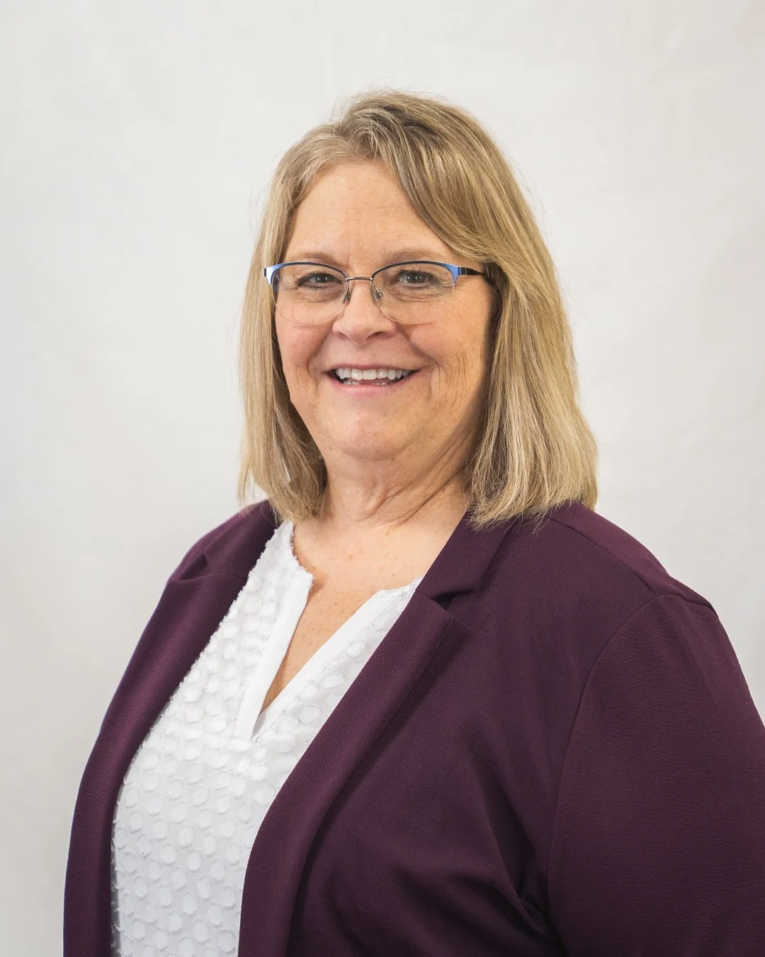 A middle-aged woman with shoulder-length blond hair, wearing glasses, a white blouse, and a dark purple blazer, smiling at the camera against a plain white background.