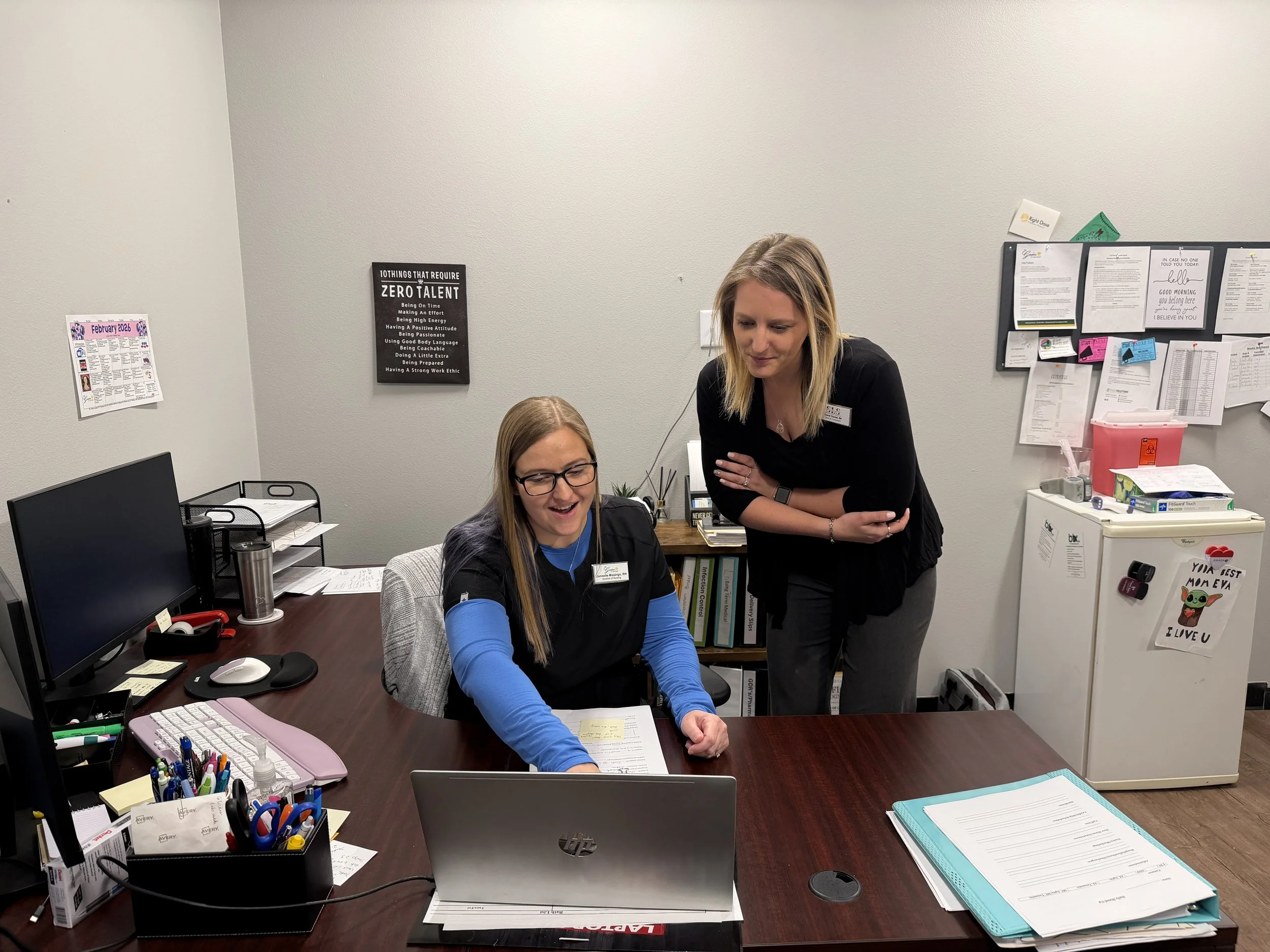 Two women working together at a desk in an office. One woman is sitting and looking at papers, while the other woman is standing and leaning over to look at the papers. The desk has two monitors, pens, and office supplies. The background has a bulletin board and framed signs on the wall.