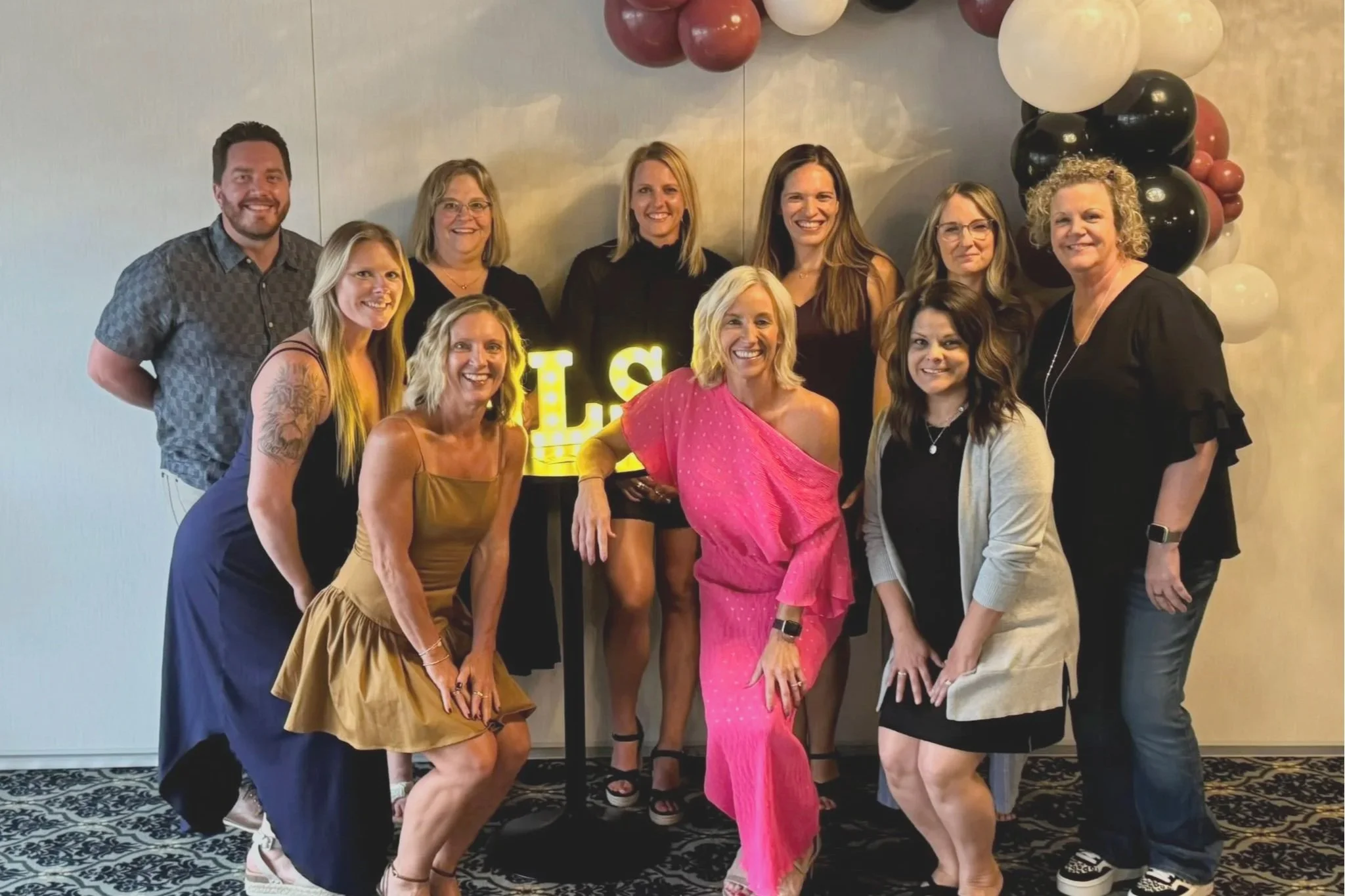 A group of eleven women and one man posing together at a celebration, with balloons and a glowing sign reading 'E' in the background. They are dressed in party attire, smiling for the photo.