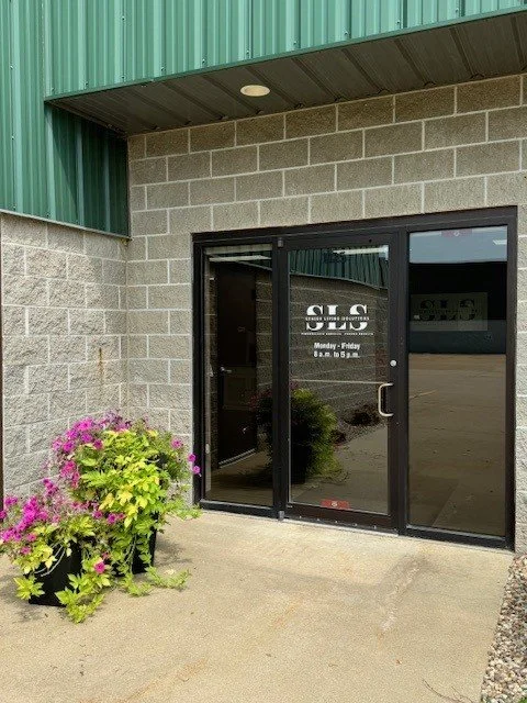 Glass door with signage, plants with pink flowers outside, building with concrete block wall and green metal roof above.