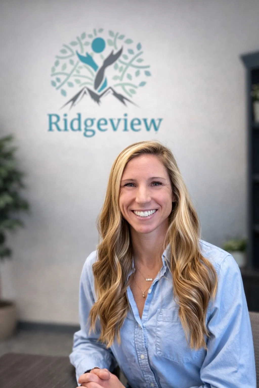 A smiling woman with long wavy blonde hair, wearing a light blue button-up shirt, sitting in a professional setting with a Ridgeview logo on the wall behind her.