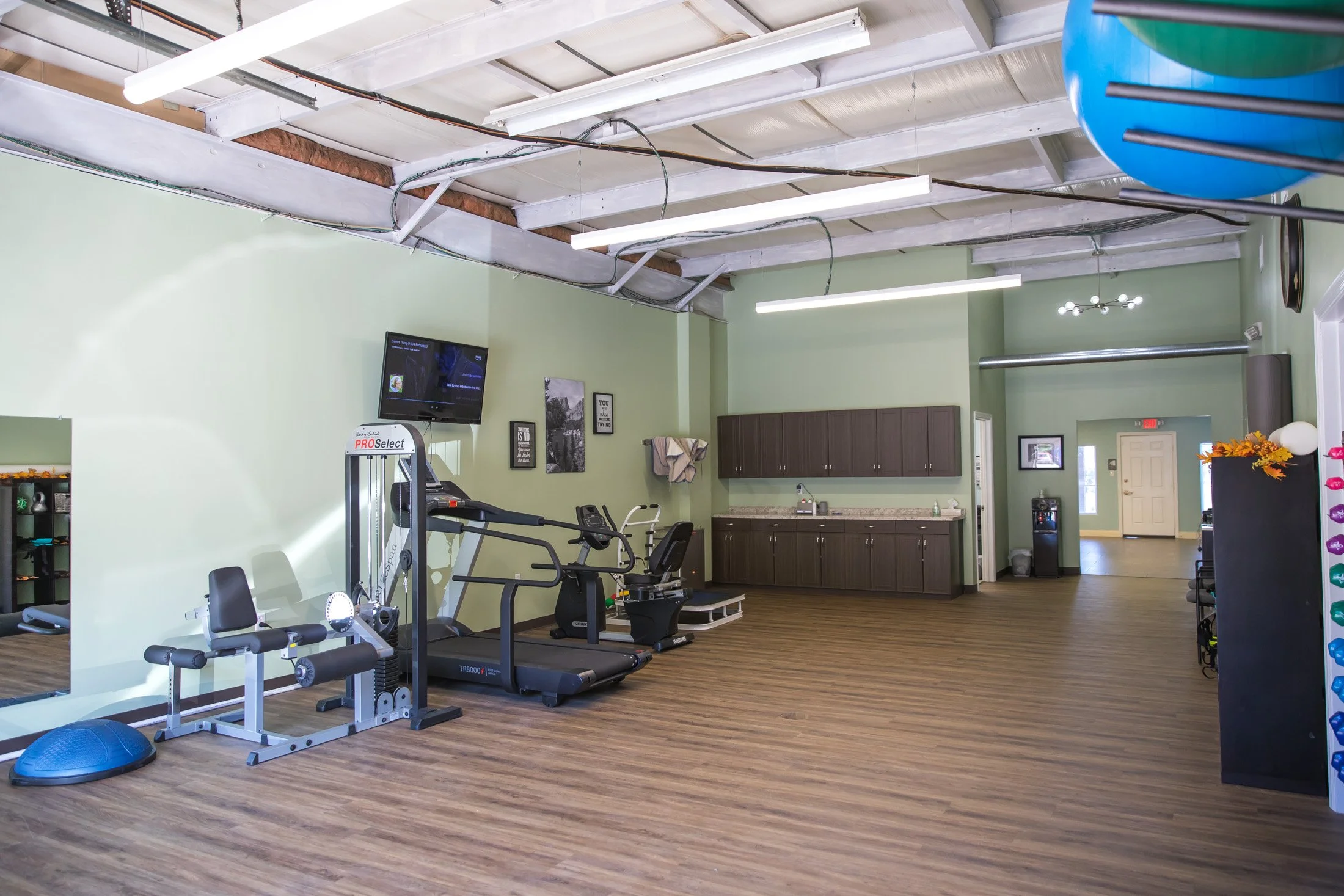 Fitness studio with workout equipment including a treadmill, exercise bike, and balance ball, a wall-mounted TV, a kitchenette area with cabinets and a sink, and a water dispenser in the corner.