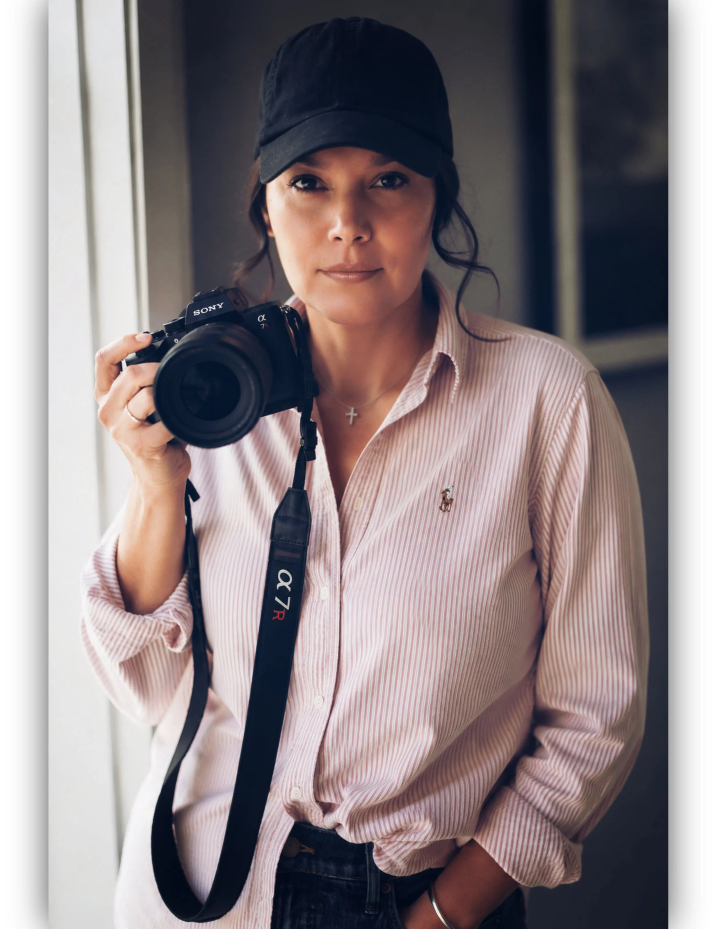 Woman wearing a black cap and pink striped shirt, holding a Sony camera, standing indoors.