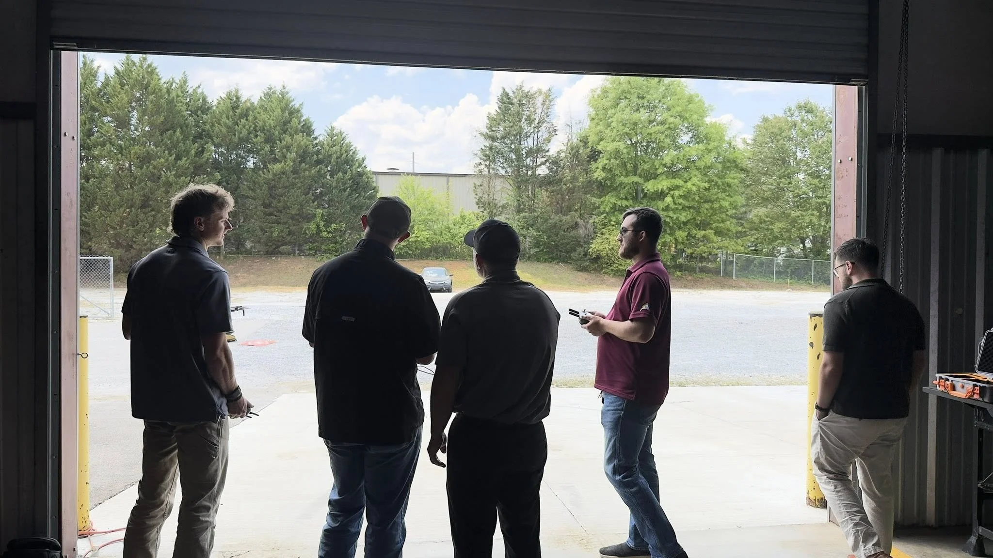 Five young men standing and talking in front of an open garage door, with a parking lot and trees visible outside.