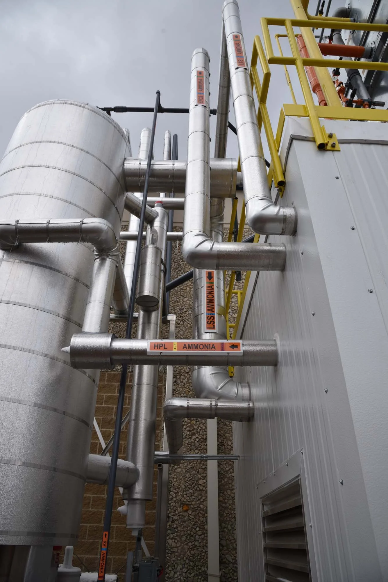 Industrial piping system with metal pipes, valves, and yellow safety railings on a building exterior, under gray sky.