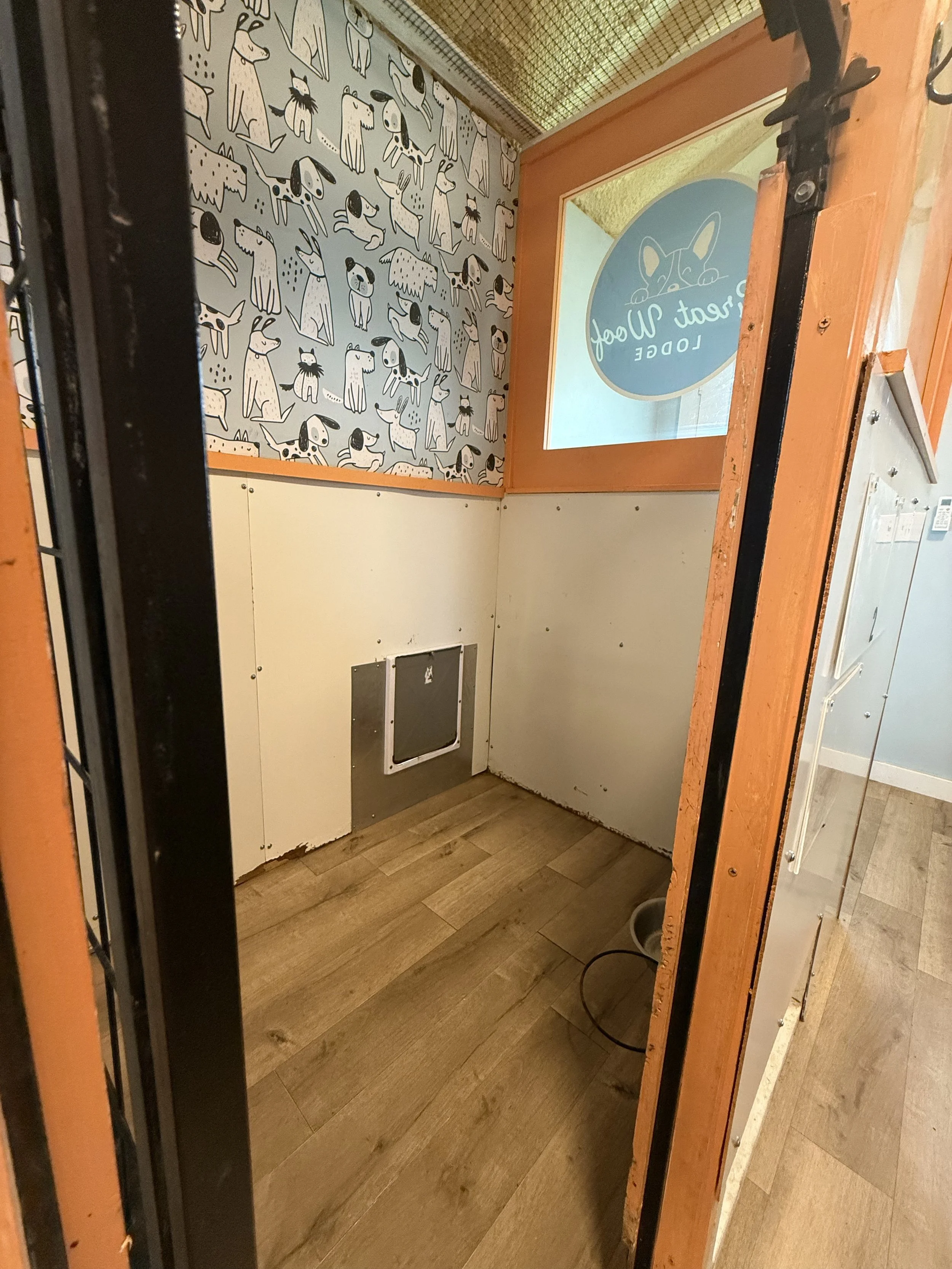 An interior view of a room with wood flooring and a partial wall with playful dog-themed wallpaper. The room appears to be under construction or renovation, with missing or incomplete wall panels and visible wiring. There is a large window or door with a sign that reads 'art walk LOOBE'.