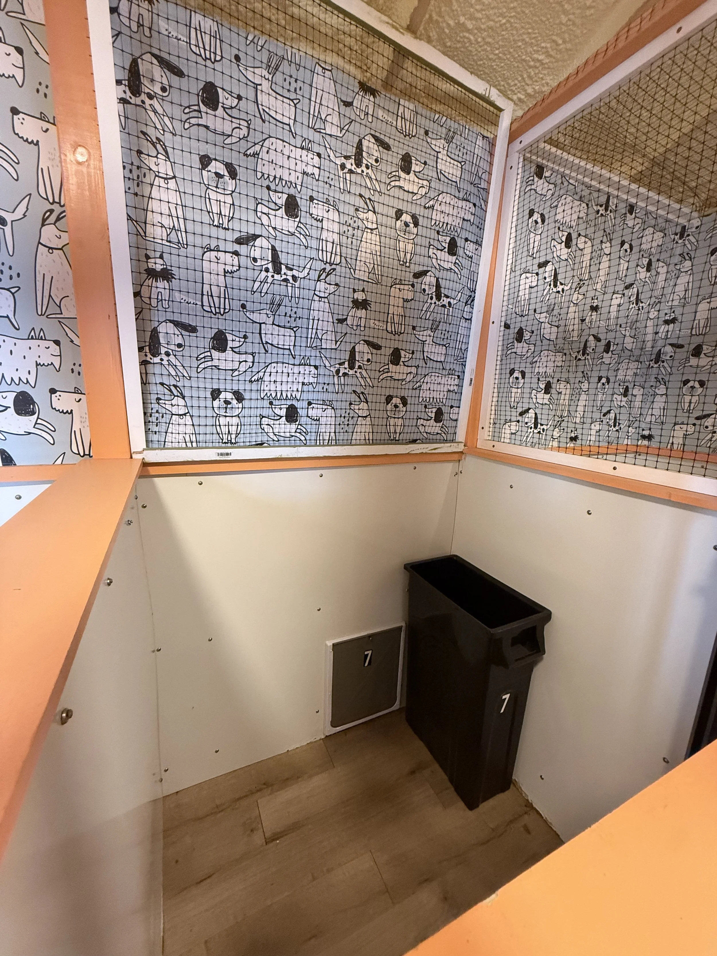 A corner of a room with a white wall decorated with a black and white dog-themed wallpaper, black bins labeled with the number 7, a black trash can, and a wooden floor.