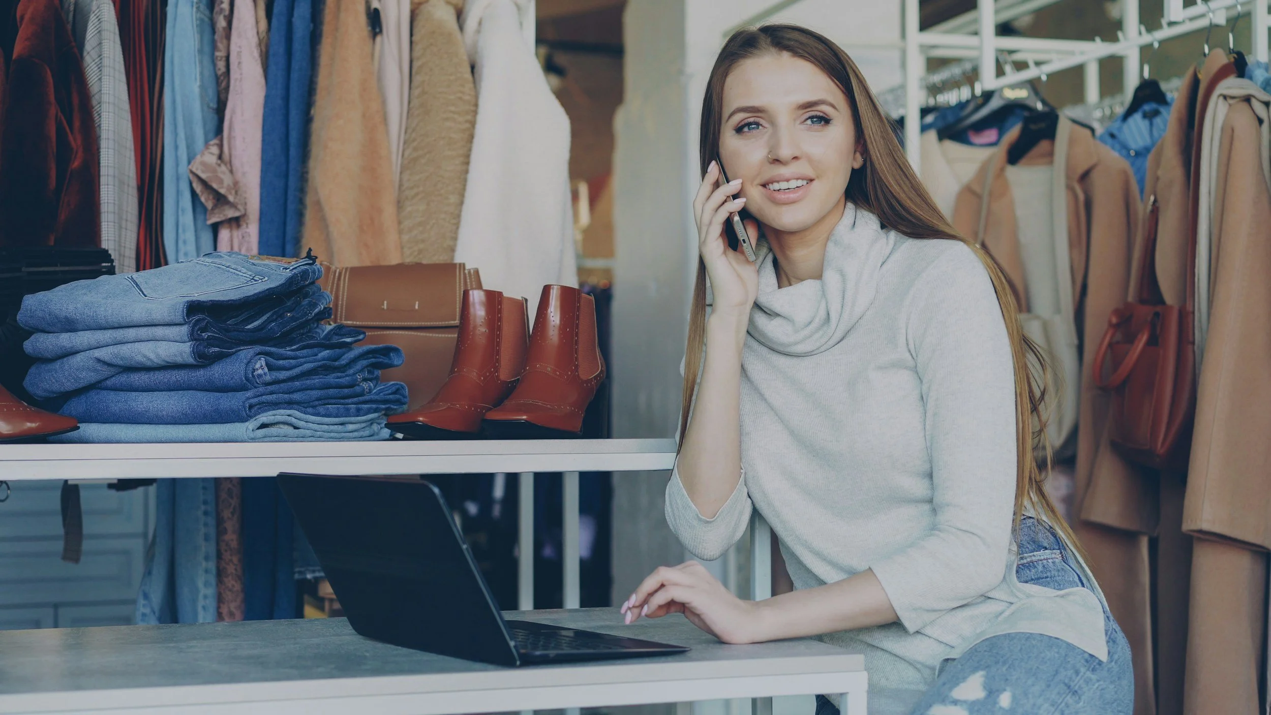 A young woman is sitting at a counter in a clothing store, talking on her cellphone. Behind her, there are racks of clothes and a shelf with folded jeans, along with a pair of brown boots on the counter.