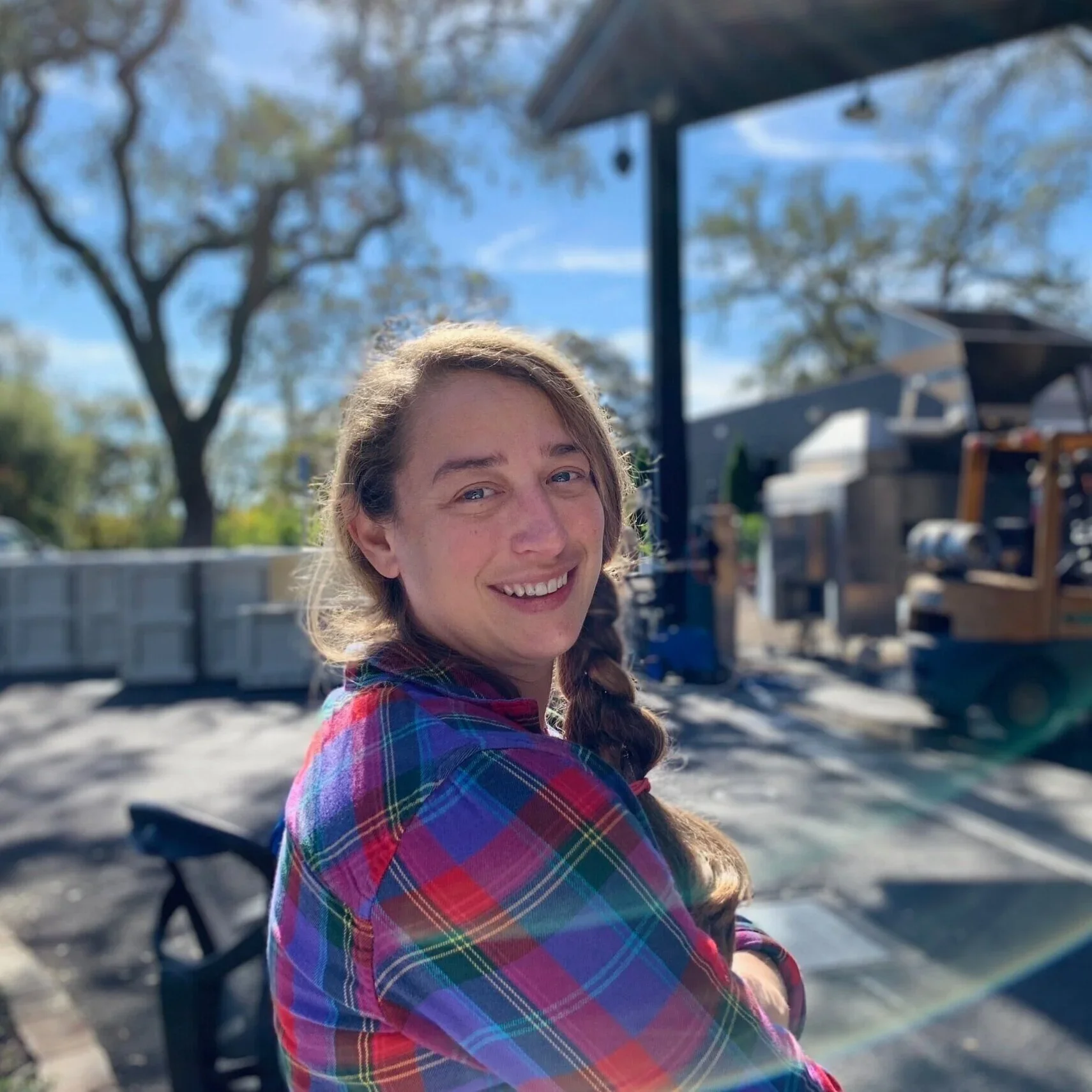 A woman with long hair wearing a plaid shirt, smiling outdoors on a sunny day with trees and a building in the background. Danielle Langlois