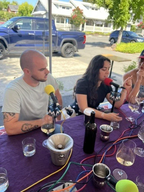 man with tattoos talks into microphone next to girl and bottles of wine