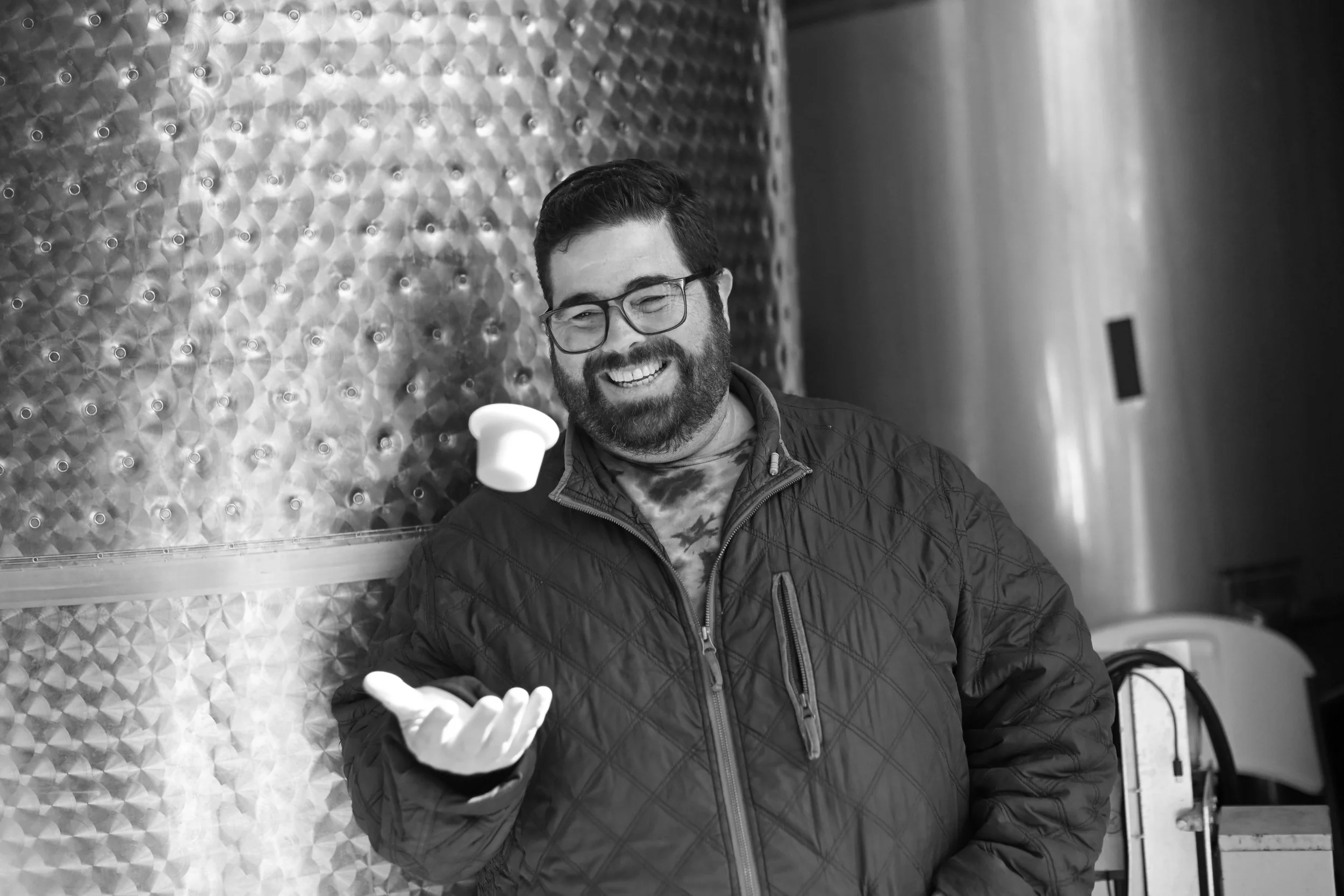 A man with glasses and a beard smiling in a black quilted jacket, gesturing with his right hand, standing in front of a textured metallic wall in an indoor setting. Sam Coturri in the winery.
