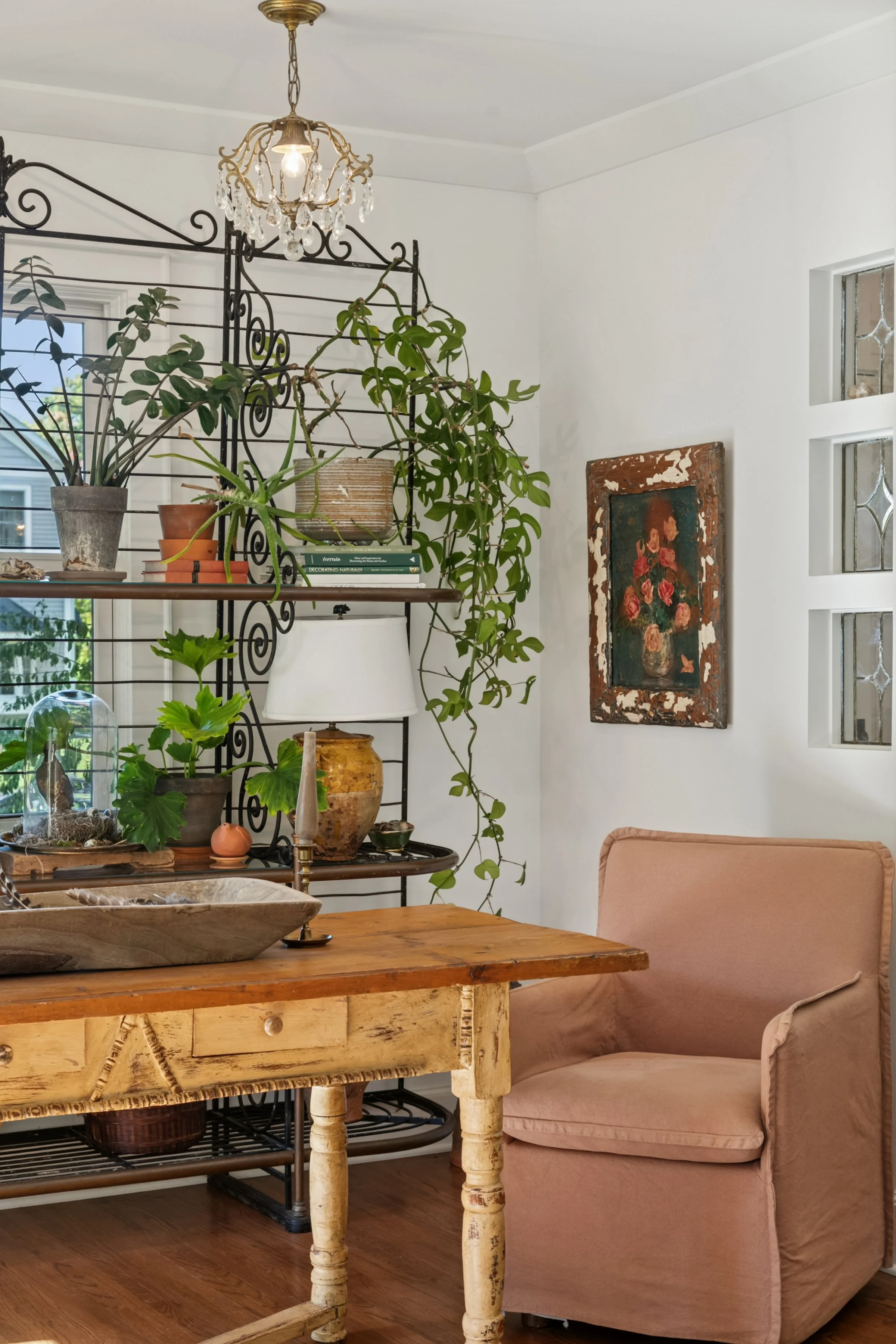 A cozy corner of a living room with a wooden table, a pink armchair, a white wall with a framed floral painting, a black metal shelving unit filled with potted plants, and a ceiling light fixture with hanging crystals.