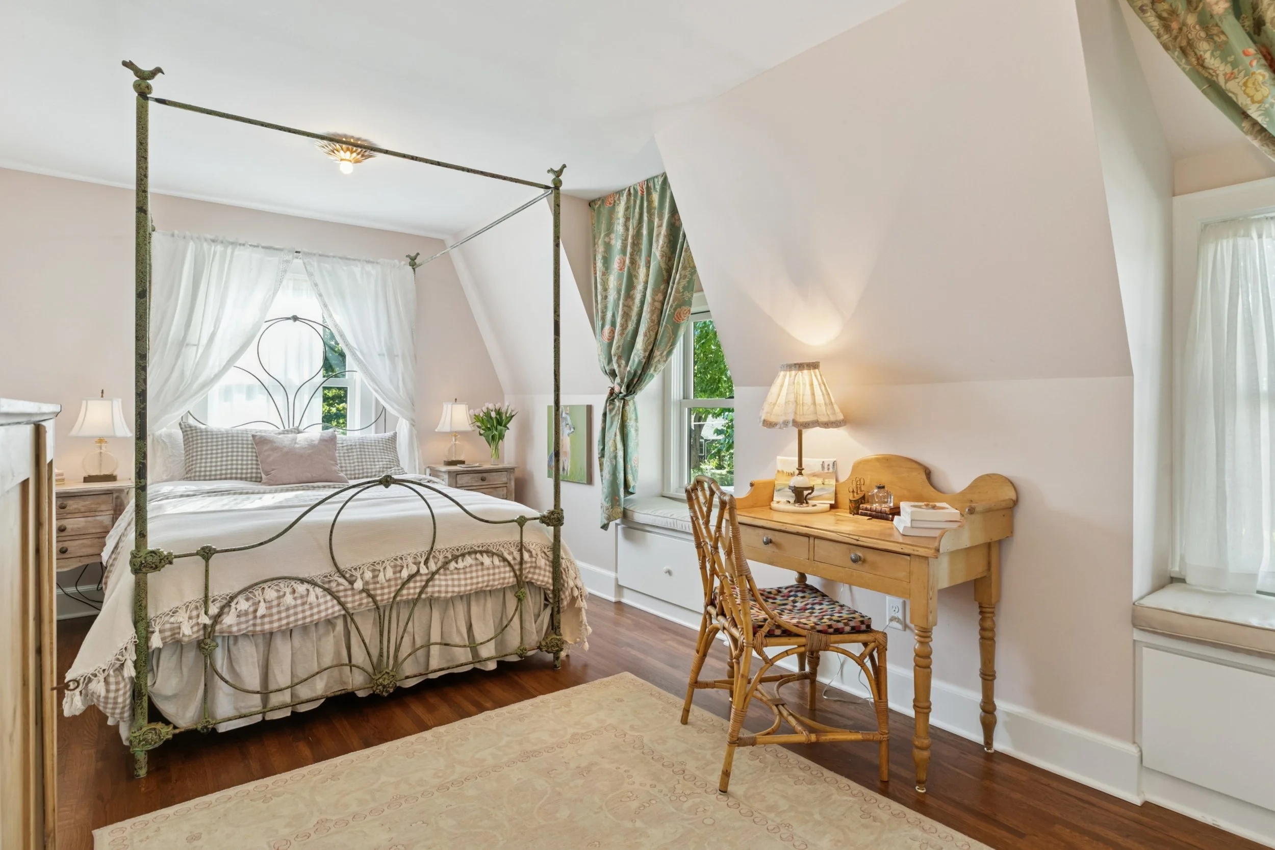 Cozy bedroom with a vintage iron canopy bed, wooden nightstands with lamps, a window with floral curtains, a wooden writing desk with books and a lamp, and hardwood floors with a rug.