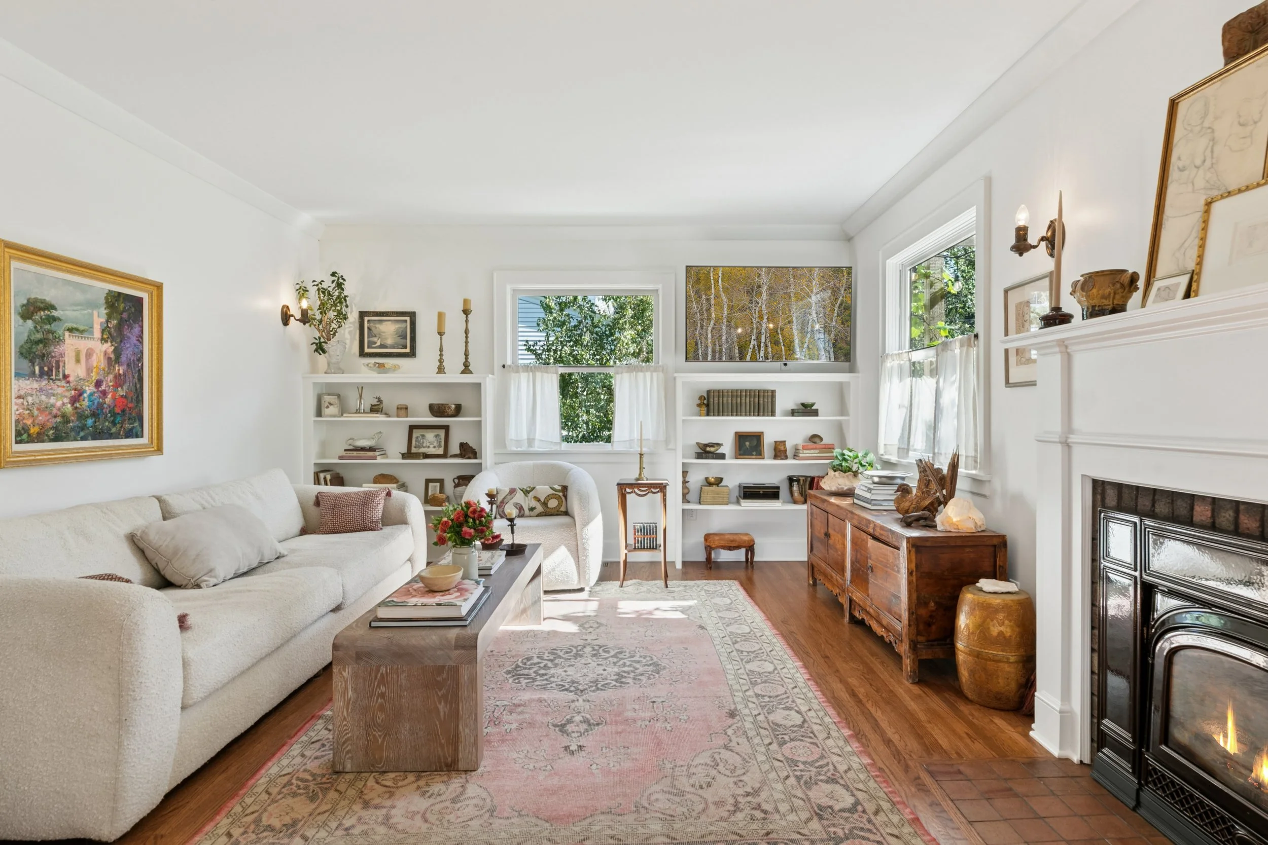 Living room with white walls, wooden floor, white sofa with pillows, wooden coffee table with books and a flower vase, bookshelf with decorations, fireplace, paintings, and windows with white curtains.